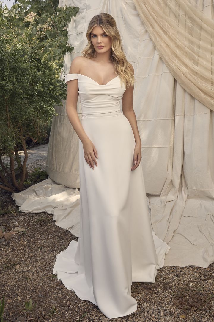 Woman in white off-the-shoulder wedding gown, standing outdoors near draped fabric and foliage.