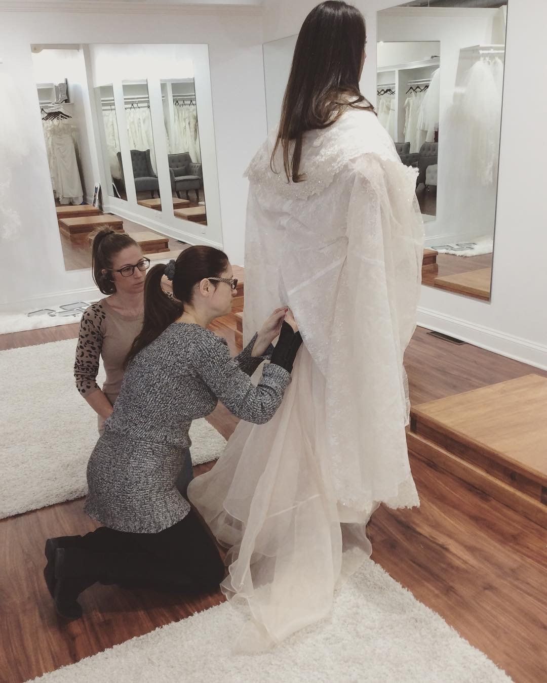 Three women in a bridal shop; two are adjusting a wedding dress on a person standing in front of a mirror.