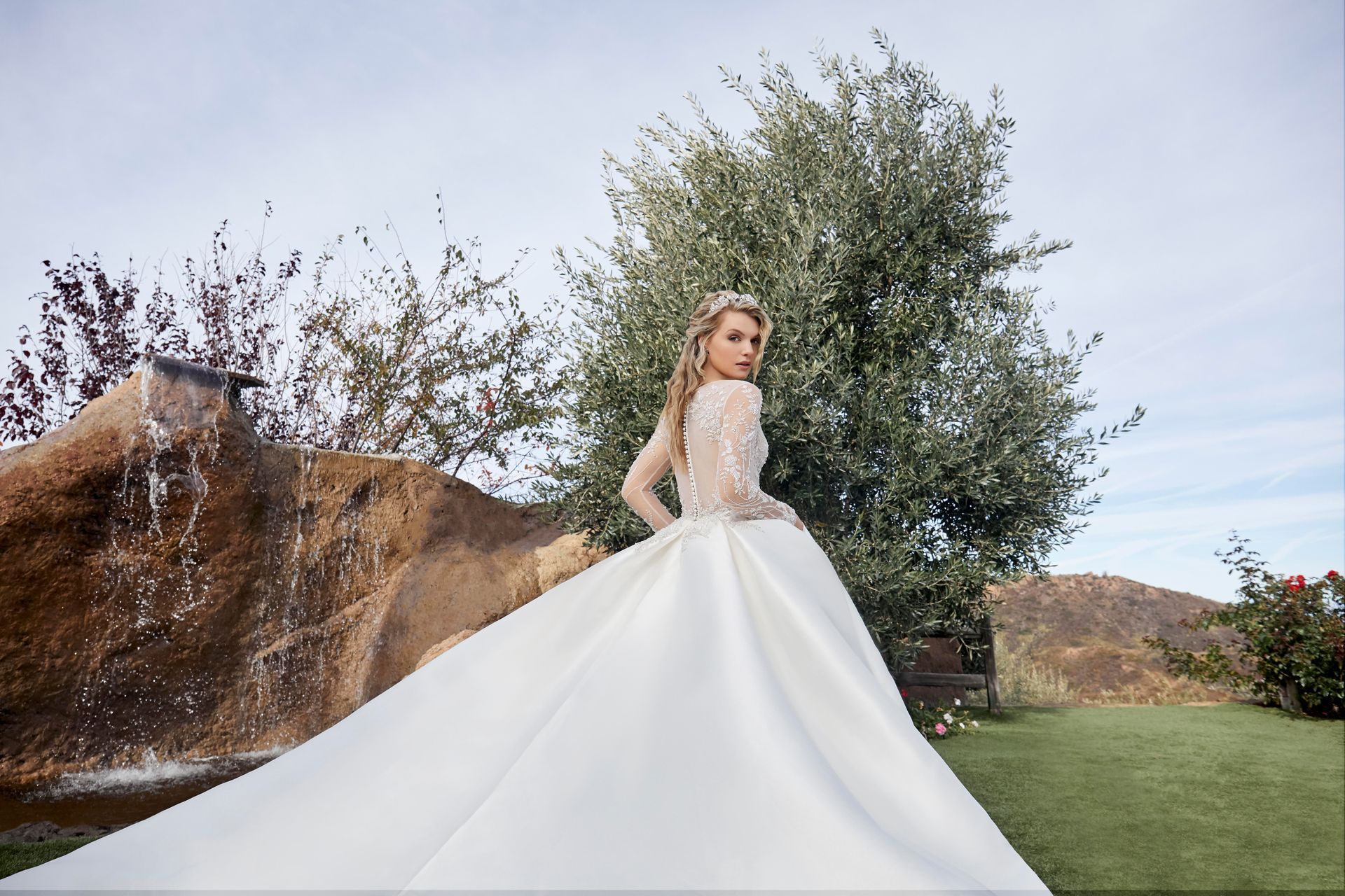 Woman in white wedding dress with a long train, standing near a waterfall, looking over her shoulder.