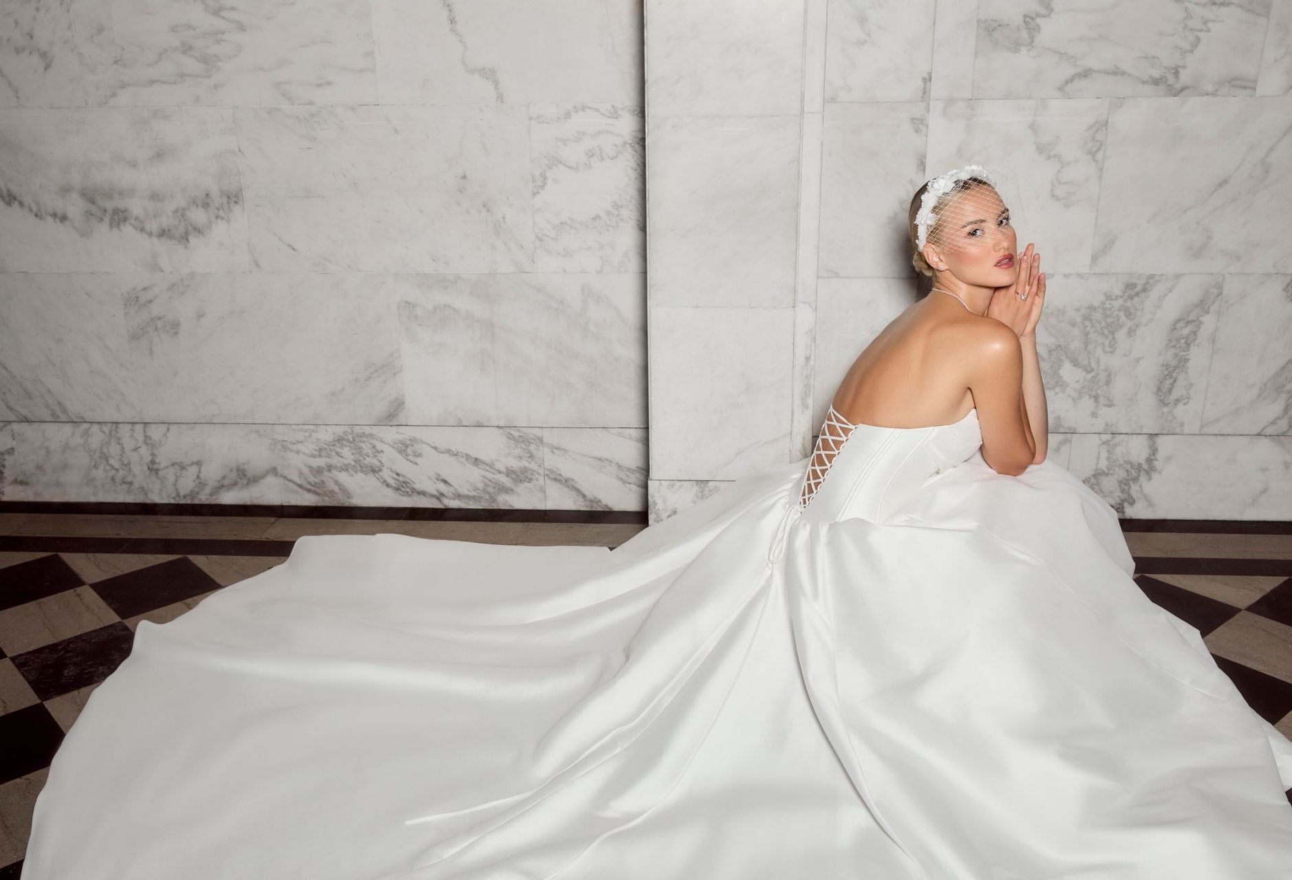 Bride in white gown with long train, sits on floor, looking over shoulder.