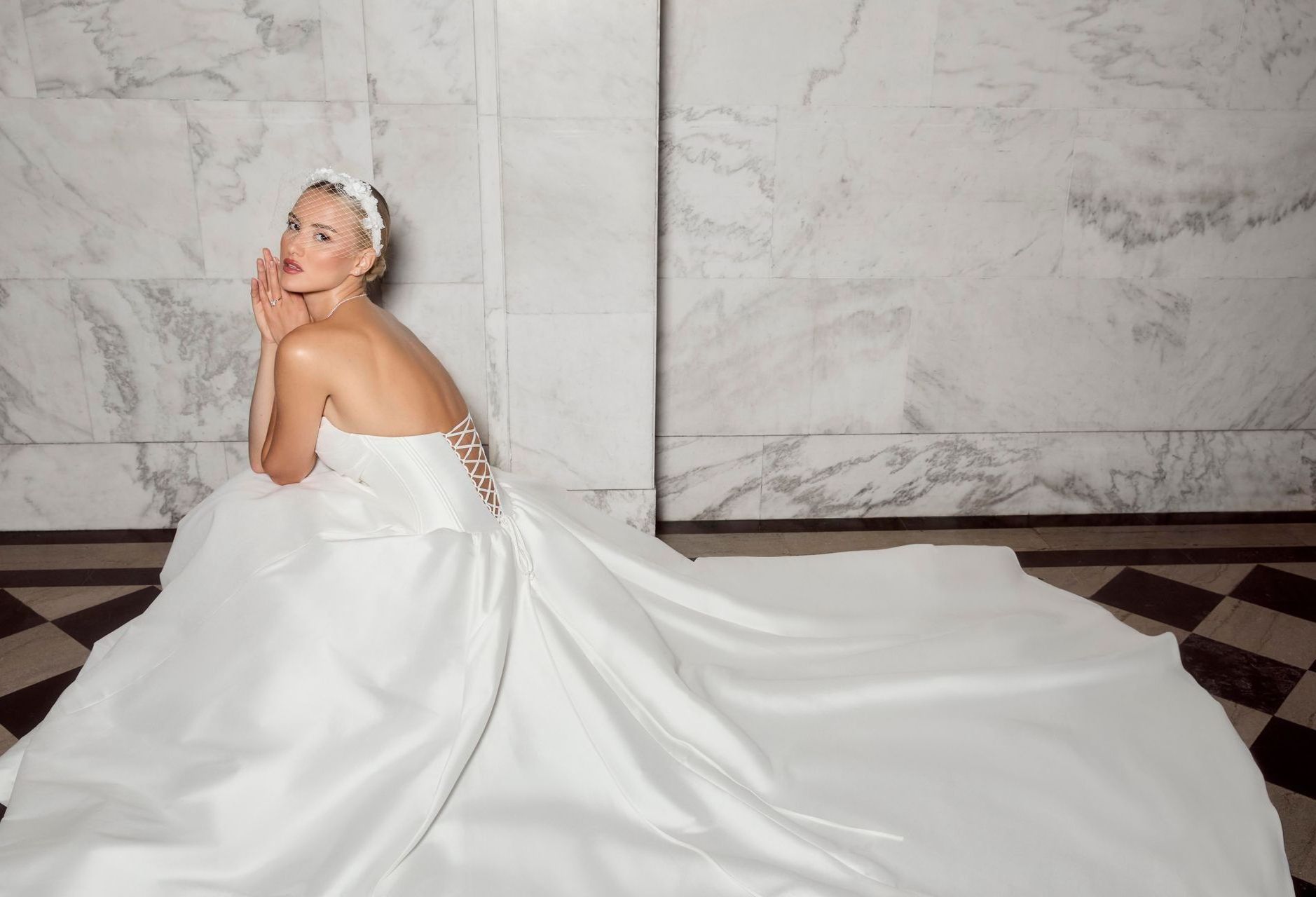 Woman in a white wedding dress, posing on checkered floor in front of marble wall.