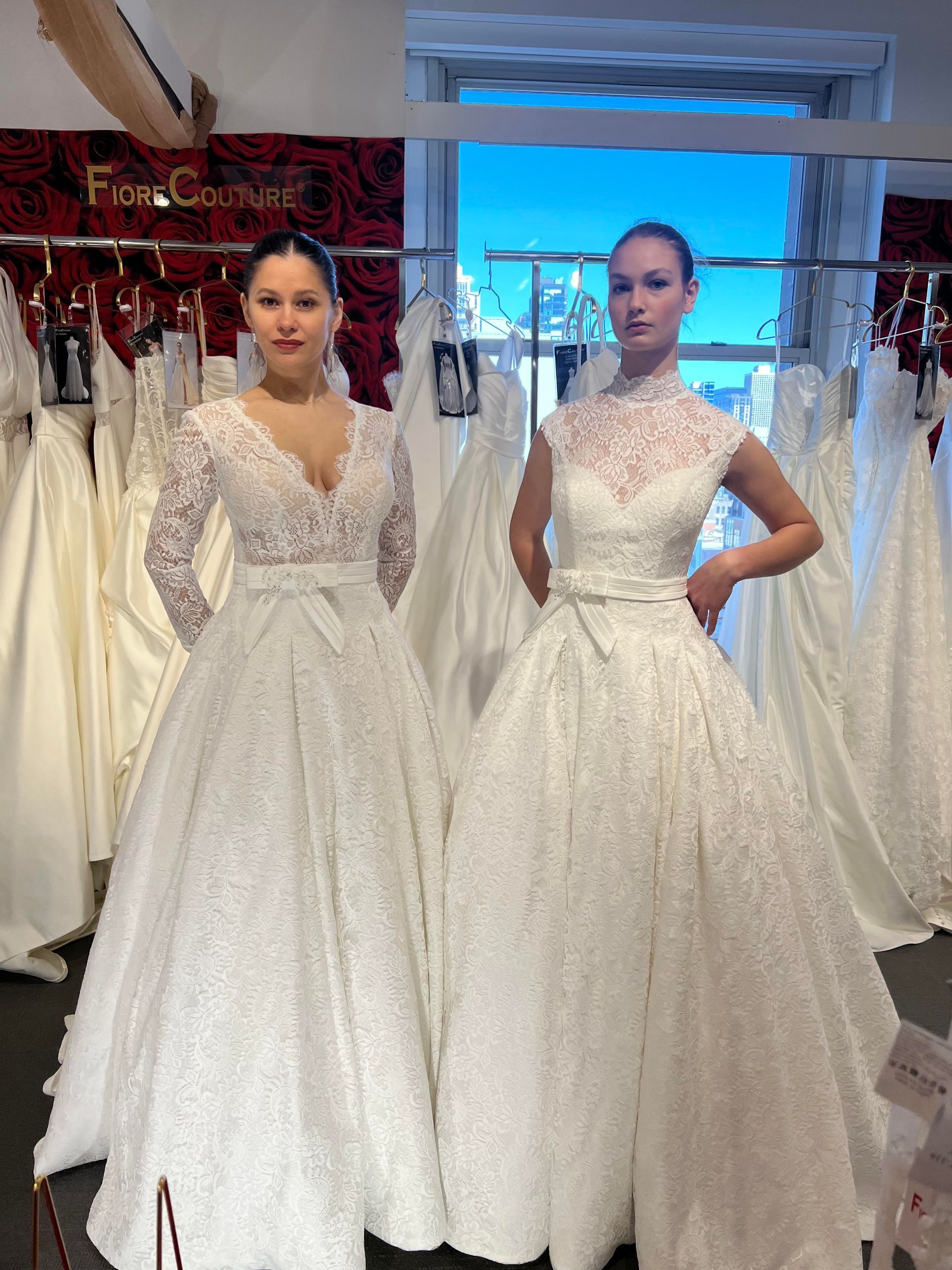 Two women in white wedding dresses, standing in a bridal shop. Dresses are embellished with lace and bows.