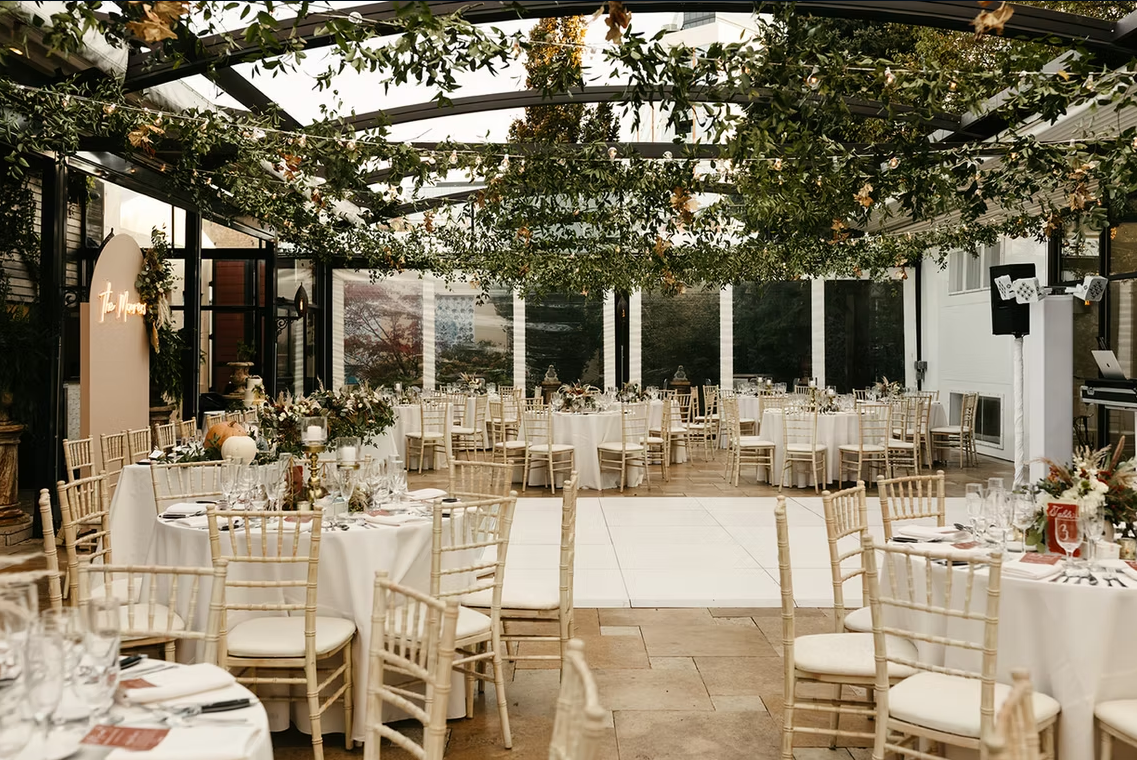 Wedding reception with round tables, decorated with flowers, under a canopy of greenery and string lights.