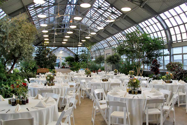 Indoor event space with round tables, white linens, and flower centerpieces, set in a greenhouse.