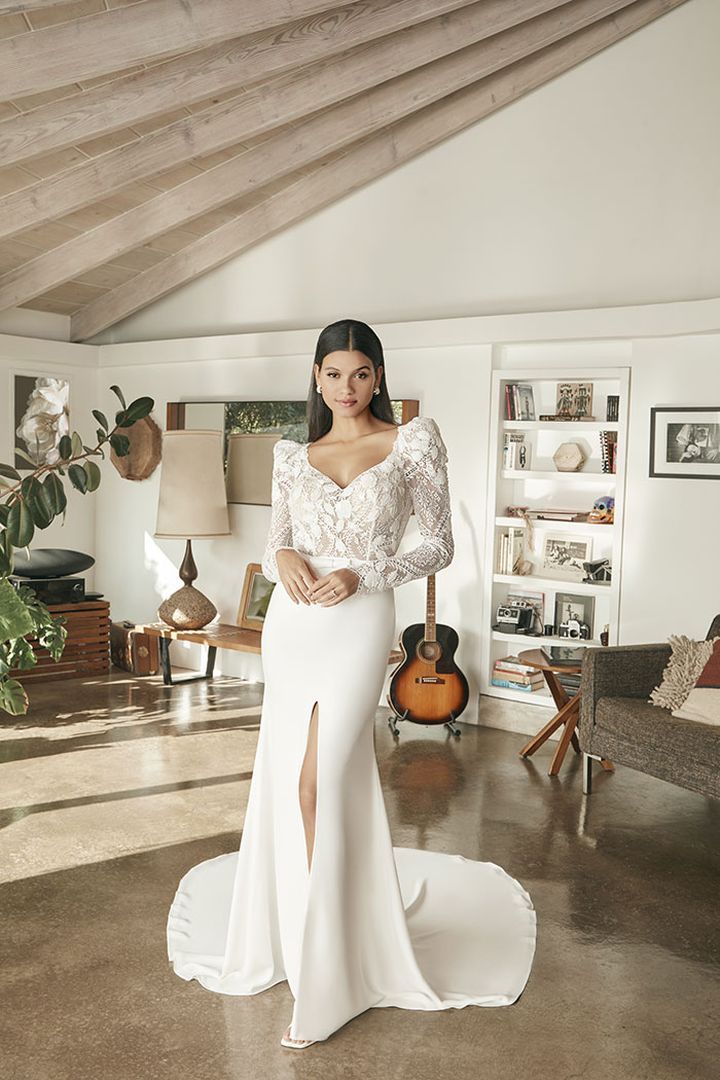 Bride in white gown with lace sleeves and a slit, standing indoors with guitar and furniture.