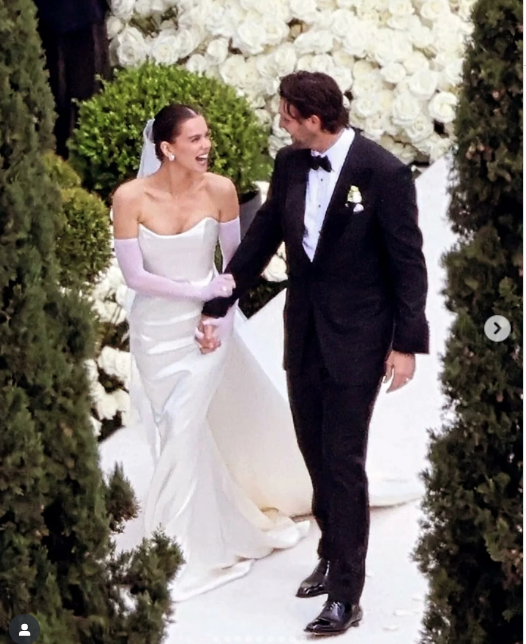 Bride and groom walking hand-in-hand. White dress, lavender gloves, black tuxedo. Outdoor setting with floral backdrop.