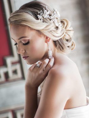 Woman with blonde updo, wearing a floral hairpiece and dangling earrings, touching her shoulder in a formal setting.