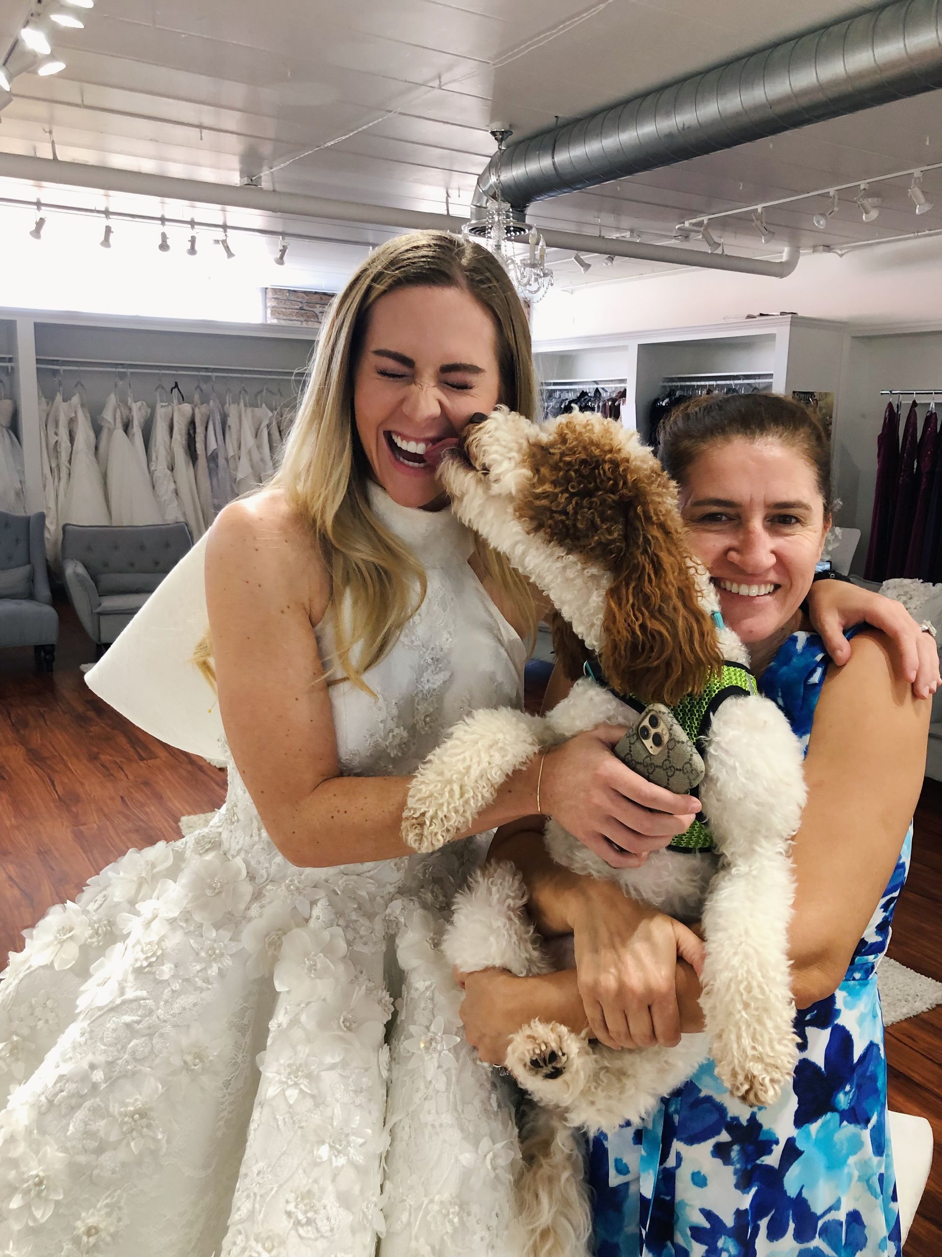 Woman in wedding dress smiles as dog licks her face; another woman hugs the dog in a bridal shop.