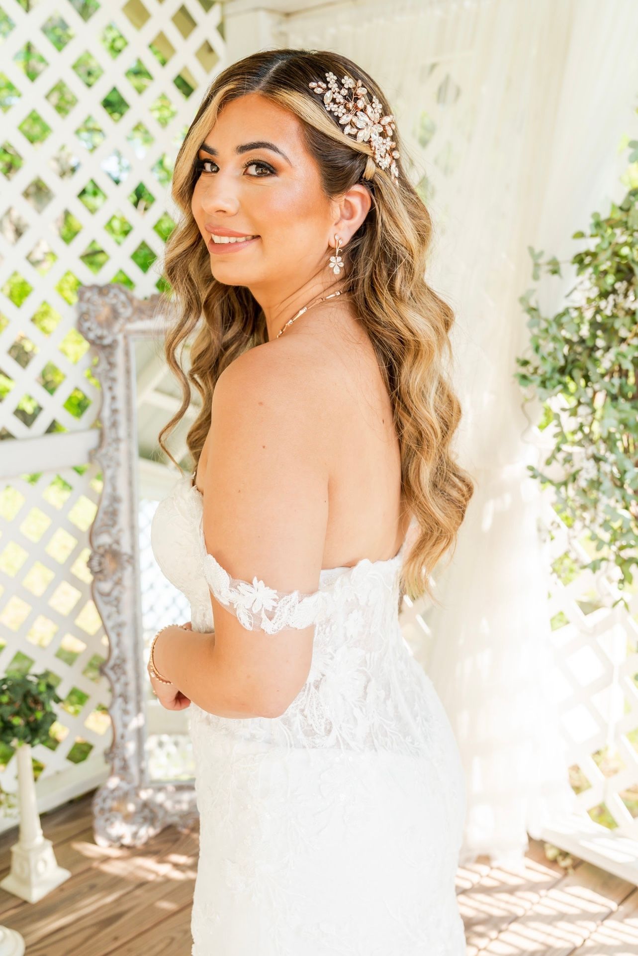Bride in a white gown smiles, gazing over shoulder. Long wavy hair, ornate headpiece, outdoor setting.