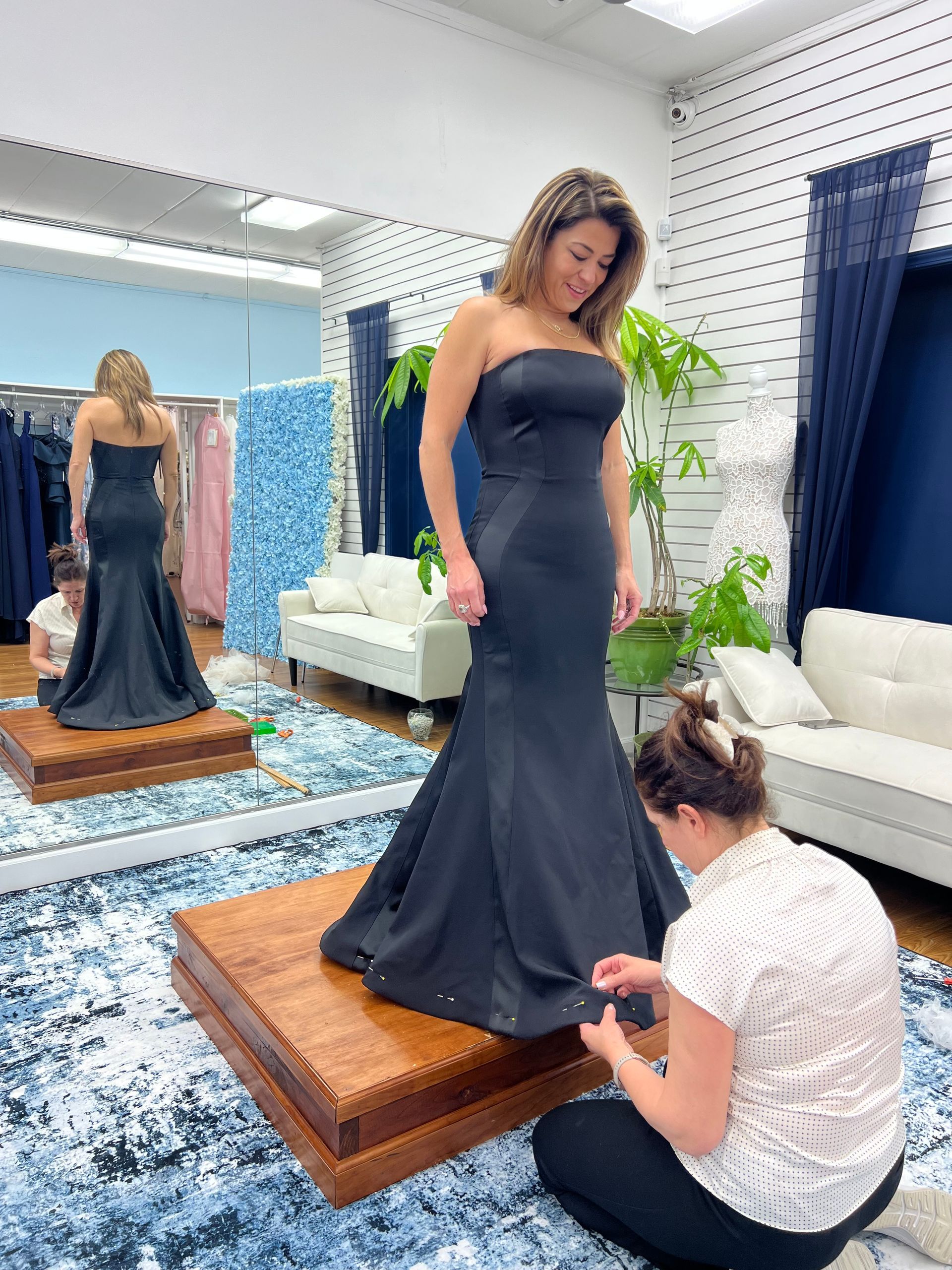 A tailor pin-fits the hem of a strapless, floor-length dark gown on a customer standing on a platform in a bridal shop.