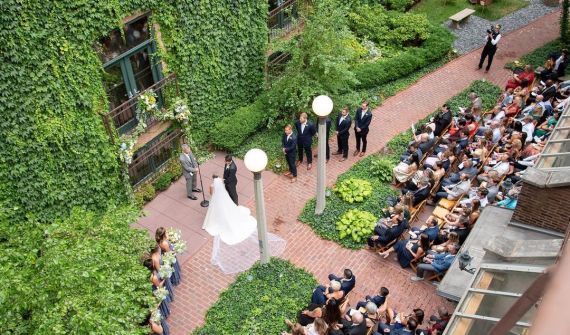 Wedding ceremony in a courtyard: bride and groom at altar, wedding party, seated guests, brick pathways, ivy-covered walls.