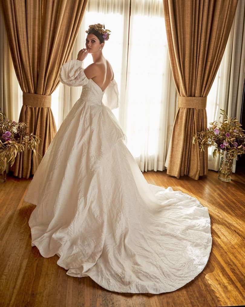 Woman in a white off-the-shoulder wedding gown turns back, standing by a window with gold curtains.