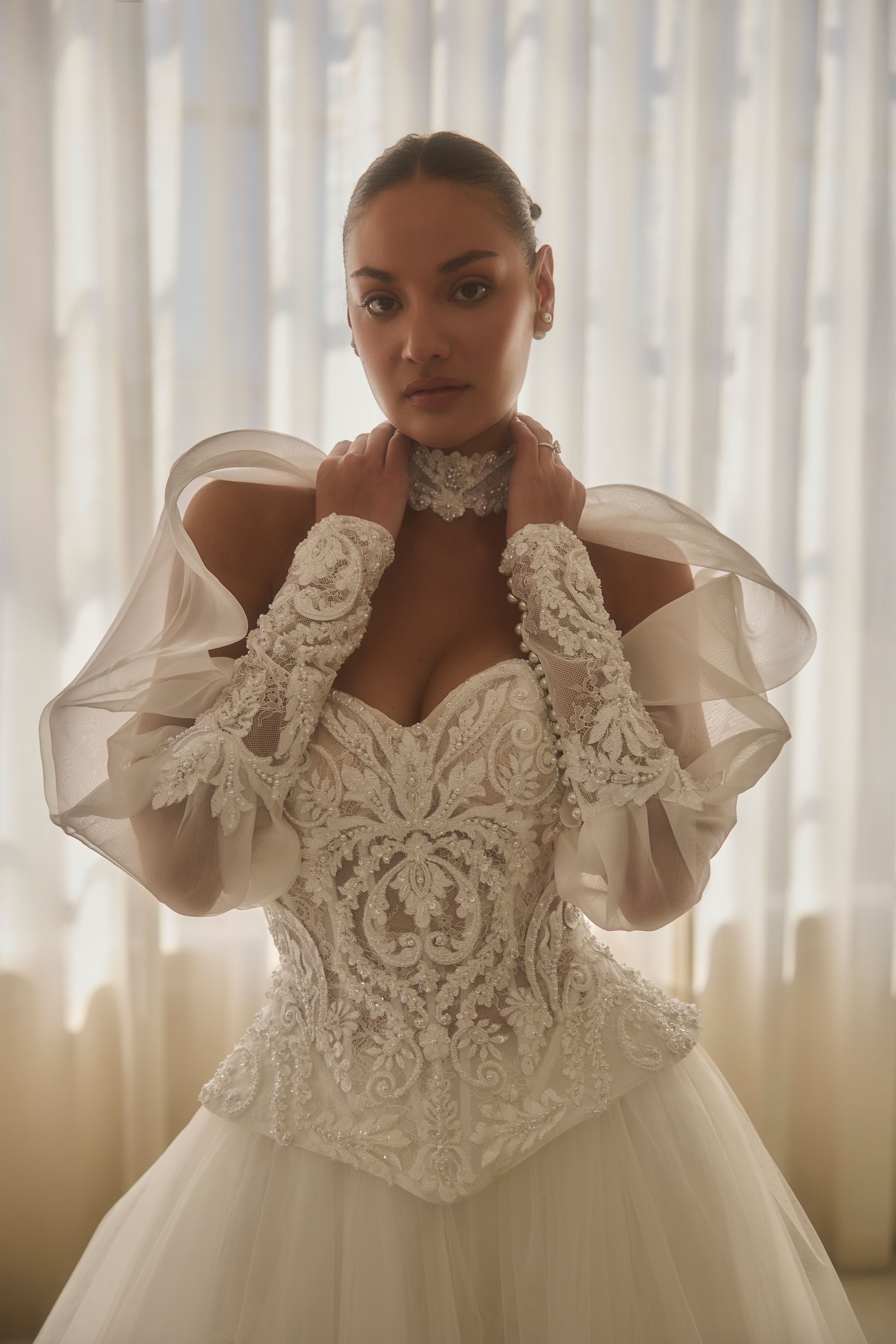Woman in a white beaded wedding dress with sheer sleeves, hands on her collar, in front of a window.