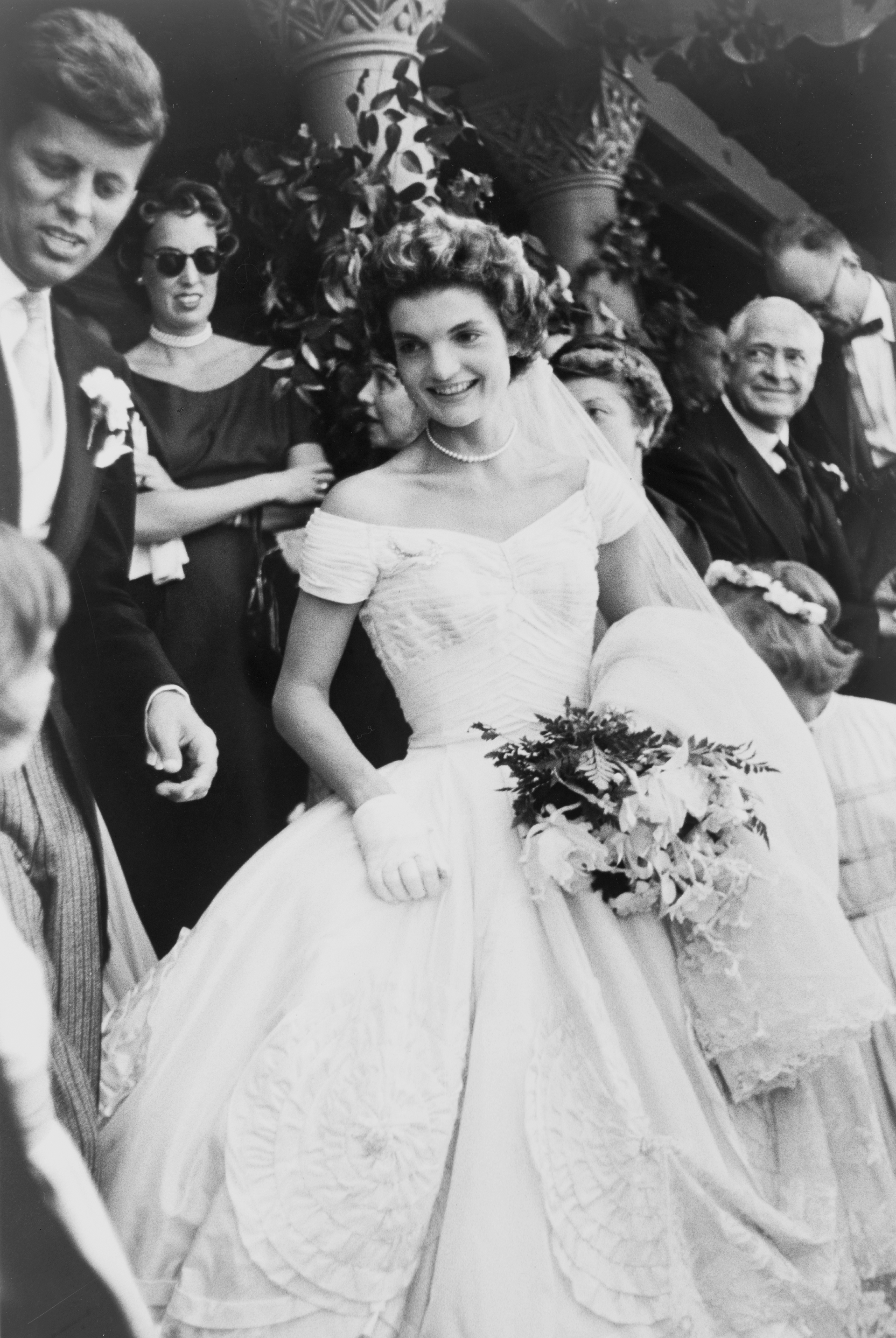 Jackie Kennedy in her wedding dress, holding a bouquet, with JFK and others nearby.