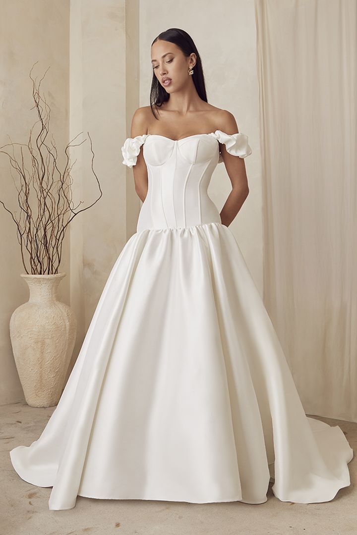 Woman in a white off-the-shoulder wedding gown. She stands indoors with a vase and neutral-toned backdrop.