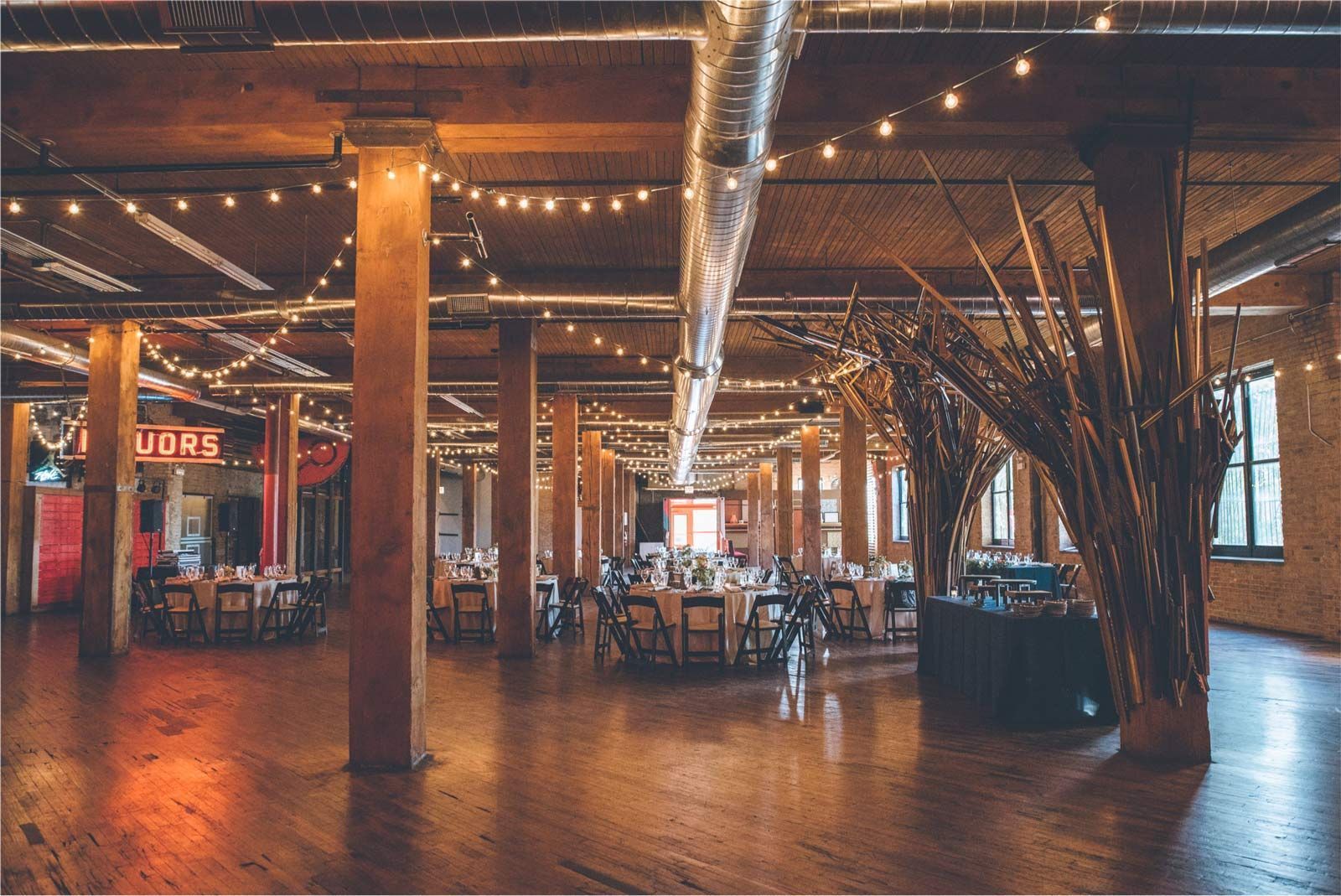 Event space with wood floors, exposed beams, string lights, round tables, and decorative branches.