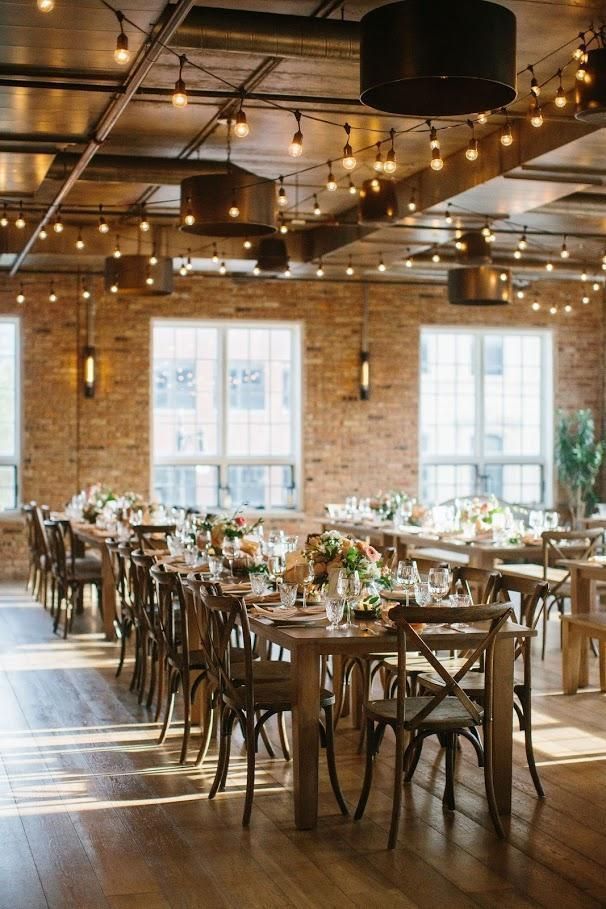 Reception hall with long wooden tables, chairs, brick walls, string lights, and large windows.