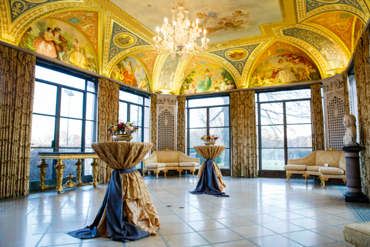 Ornate room with painted ceiling, tall windows, tables with draped cloth, and chandeliers.