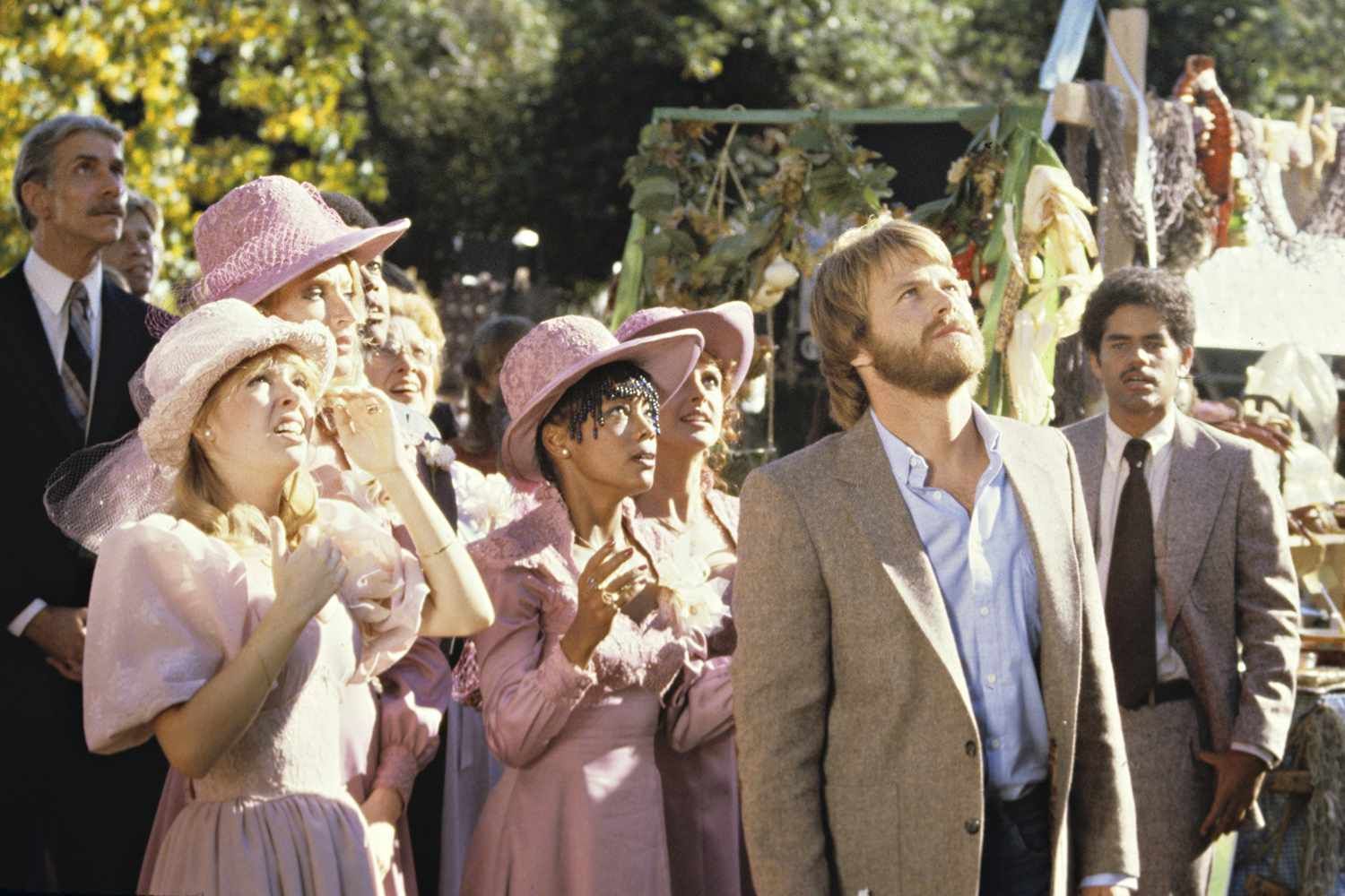 Wedding guests look upwards at an outdoor ceremony; several wear pink hats and dresses.