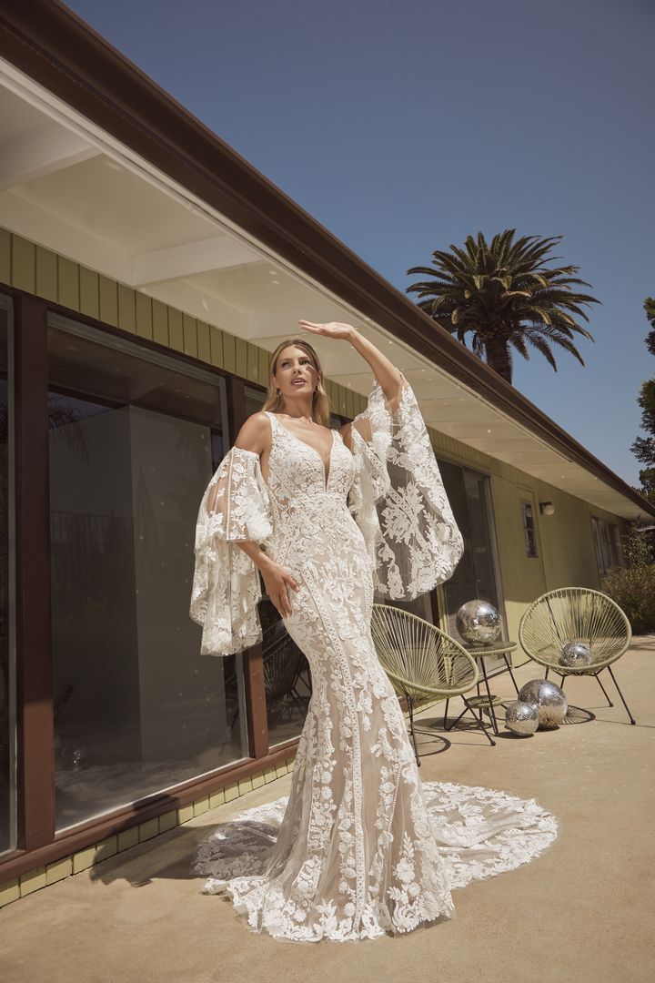 Woman in white floral wedding dress poses outside, arm raised, sunny setting.