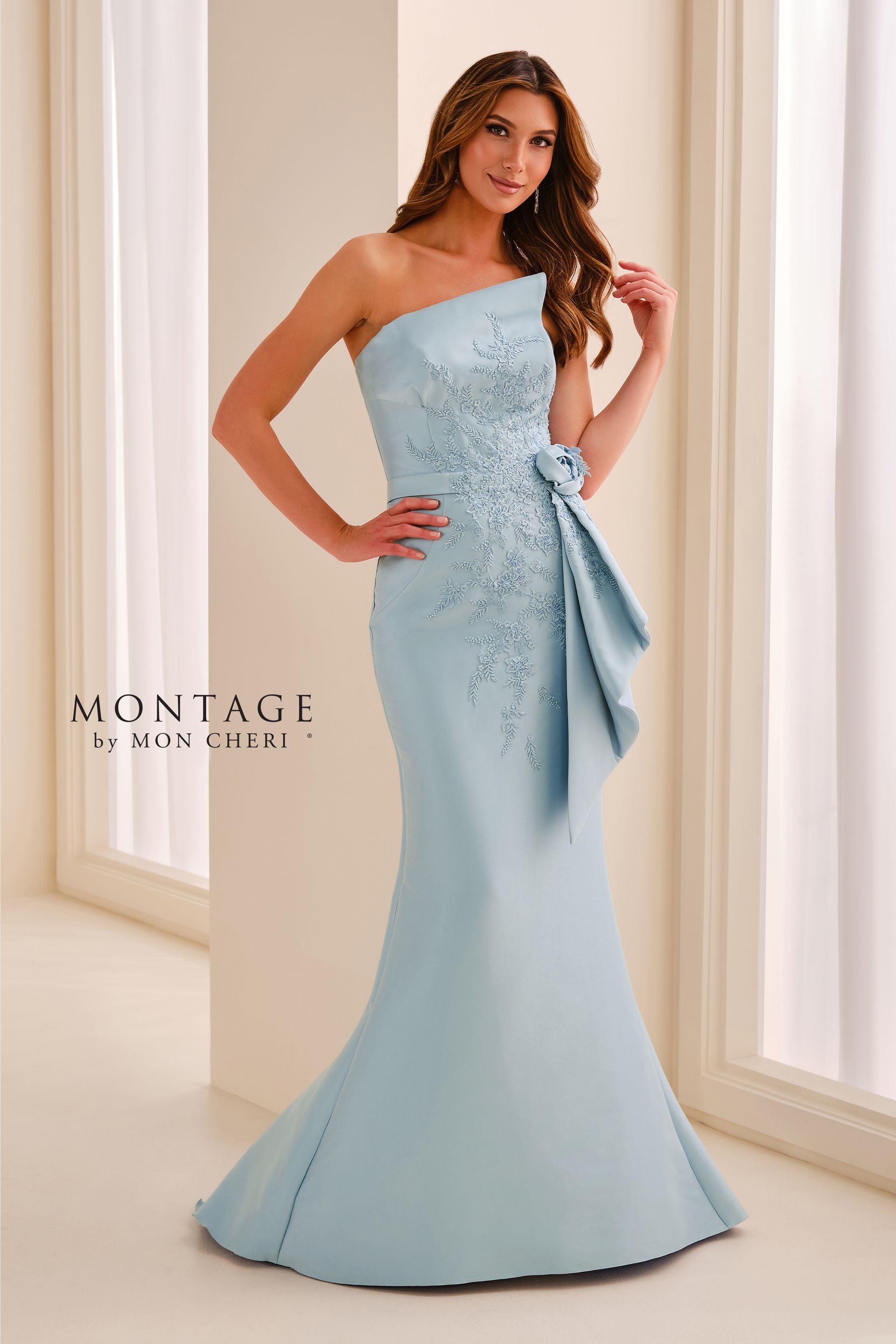 Woman in a light blue one-shoulder gown posing indoors near tall windows.