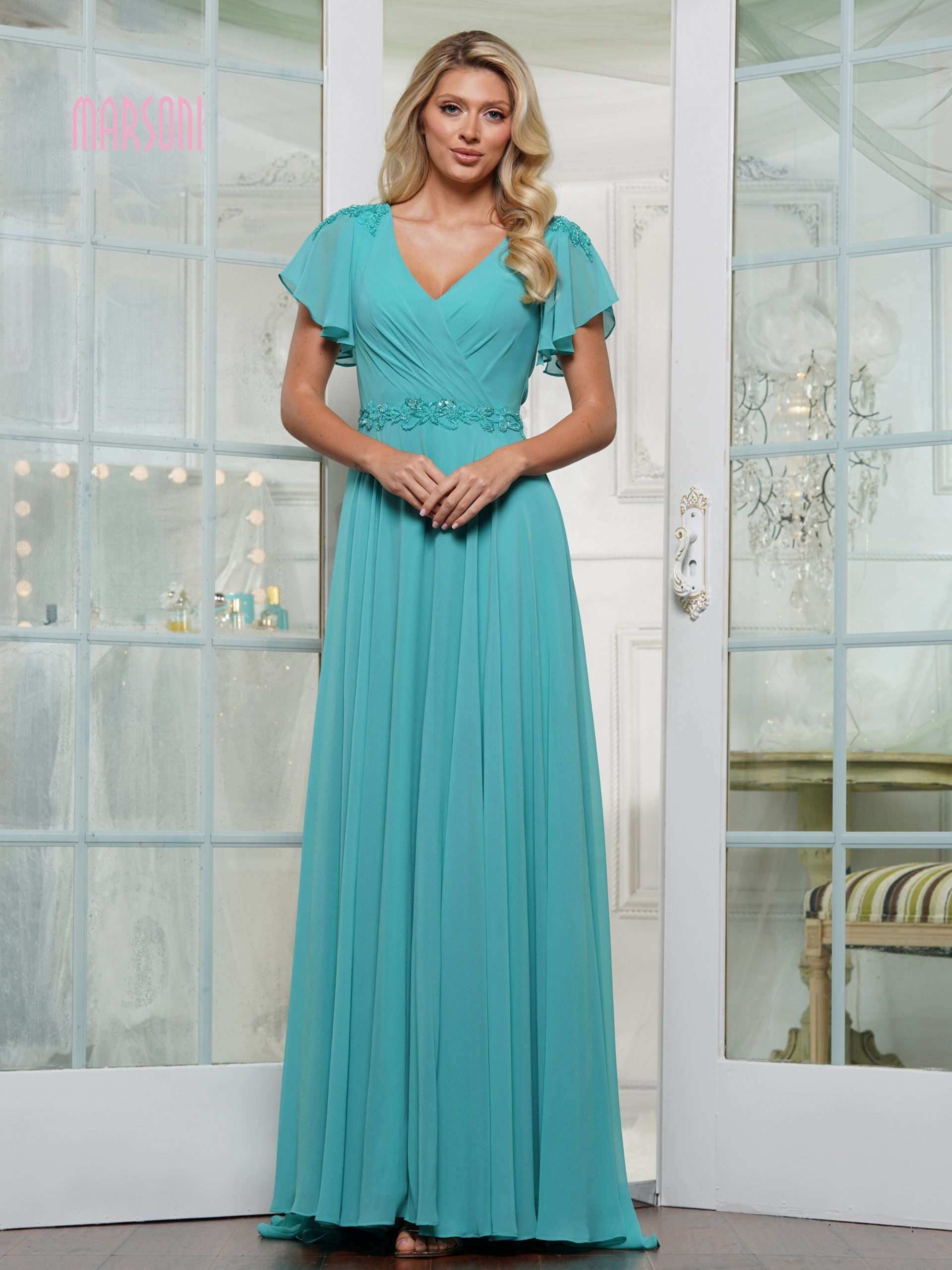 Woman in a teal chiffon gown with flutter sleeves, standing in front of white doors.