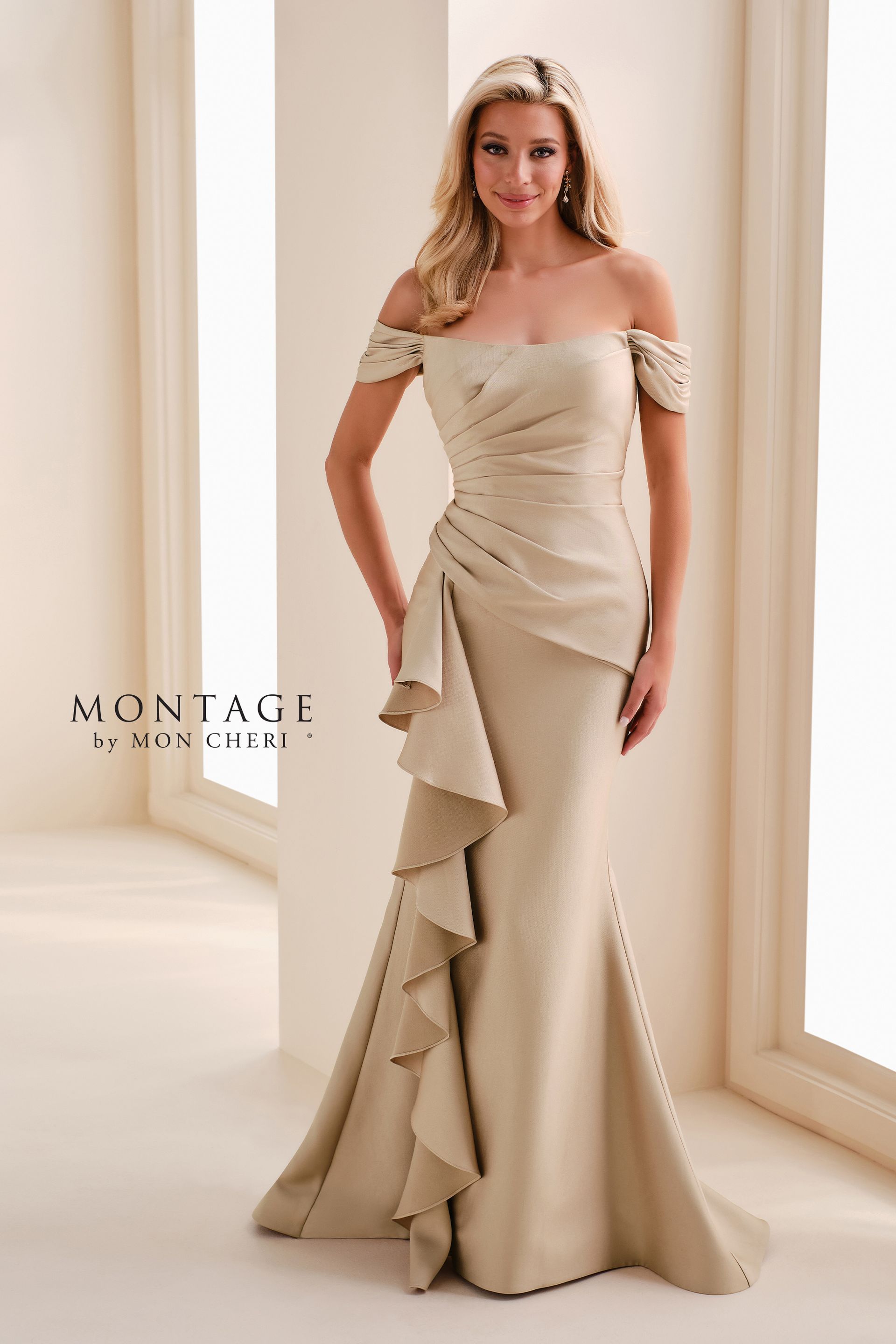 Woman in a beige off-shoulder gown with ruffled train, standing in a bright room by tall windows.