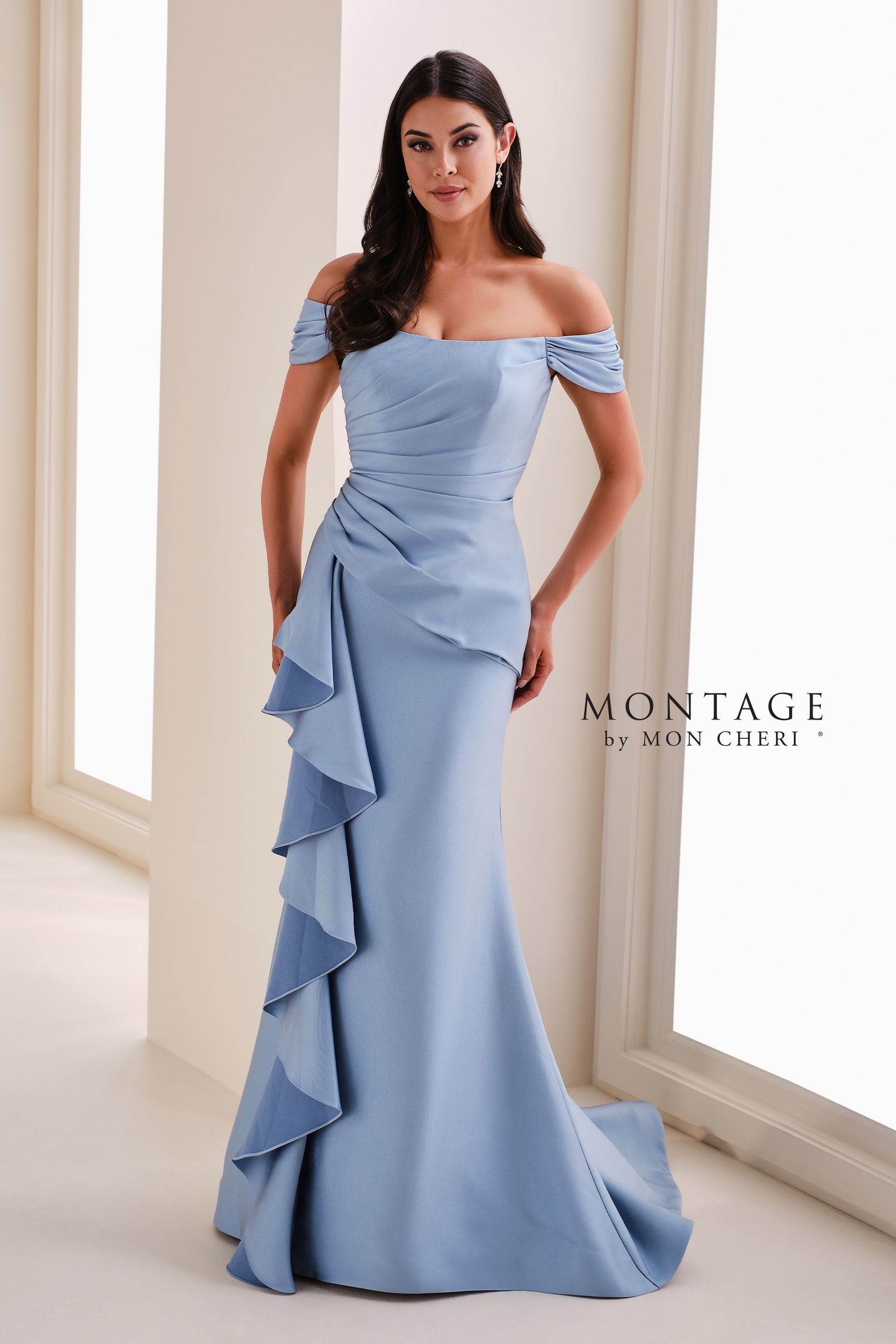 Woman in a beige off-shoulder gown with ruffled train, standing in a bright room by tall windows.