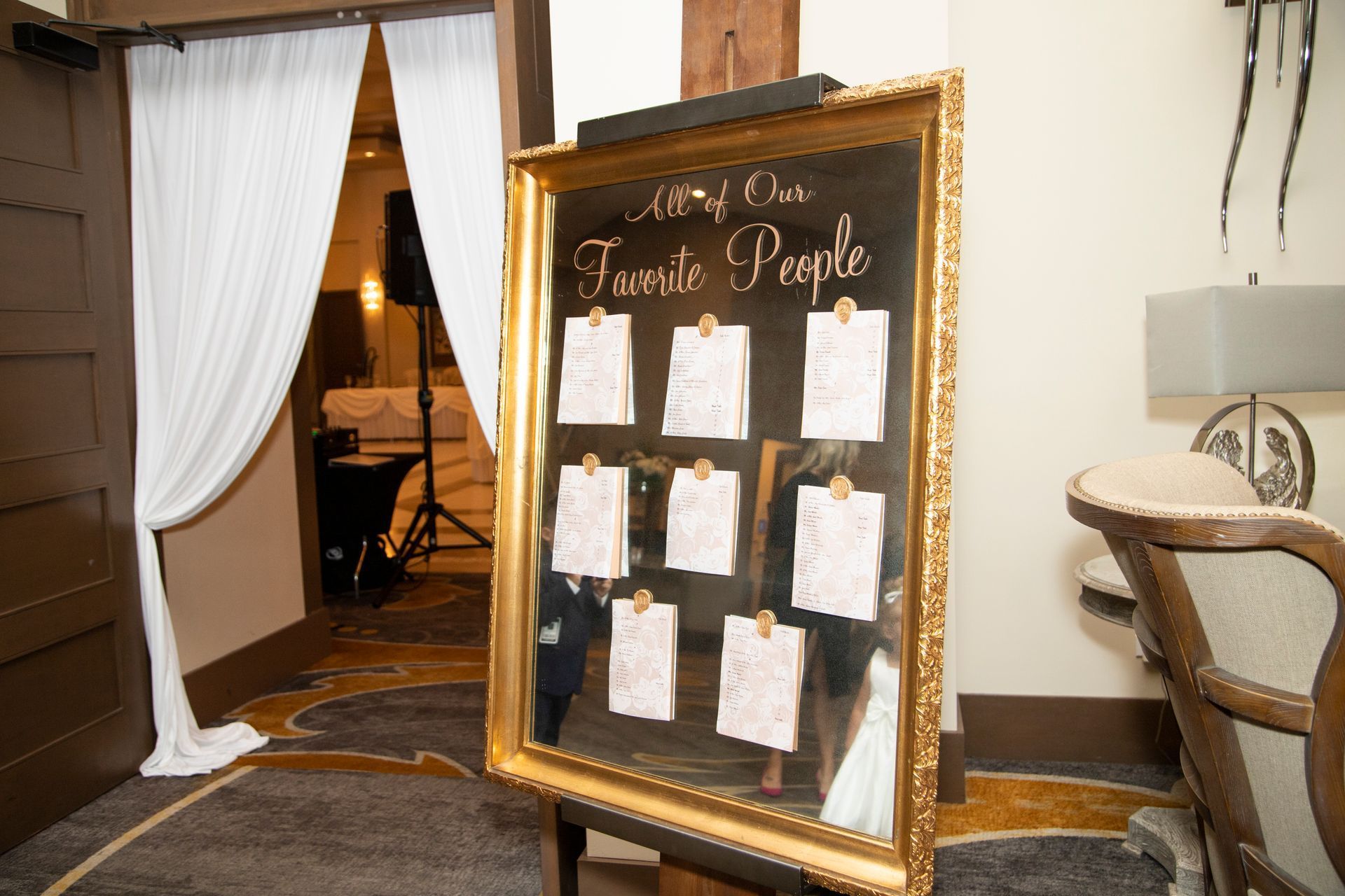 Gold-framed mirror with guest seating cards, propped on a black easel, next to a doorway with white curtains.
