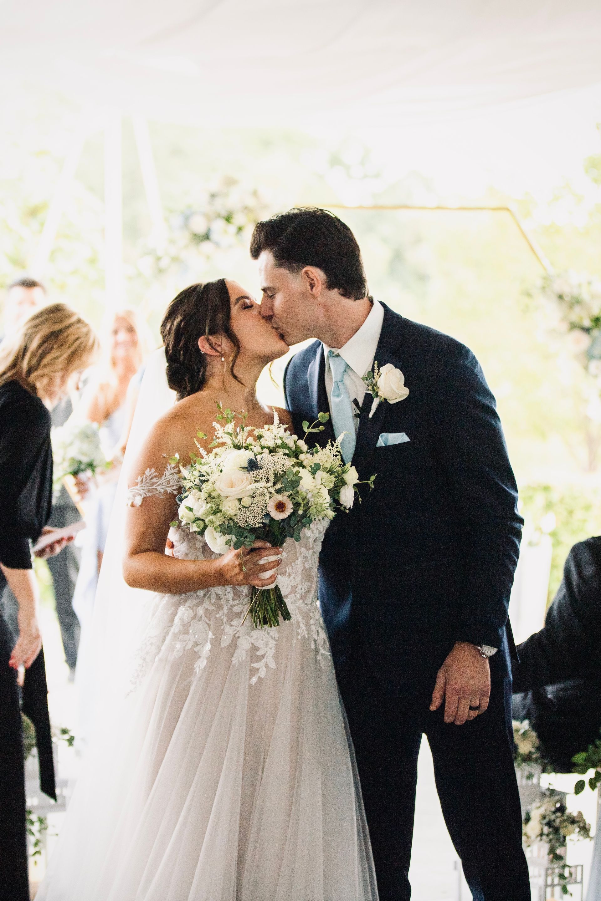 Newlyweds share a kiss during an outdoor wedding ceremony; bride in white gown, groom in a blue suit.
