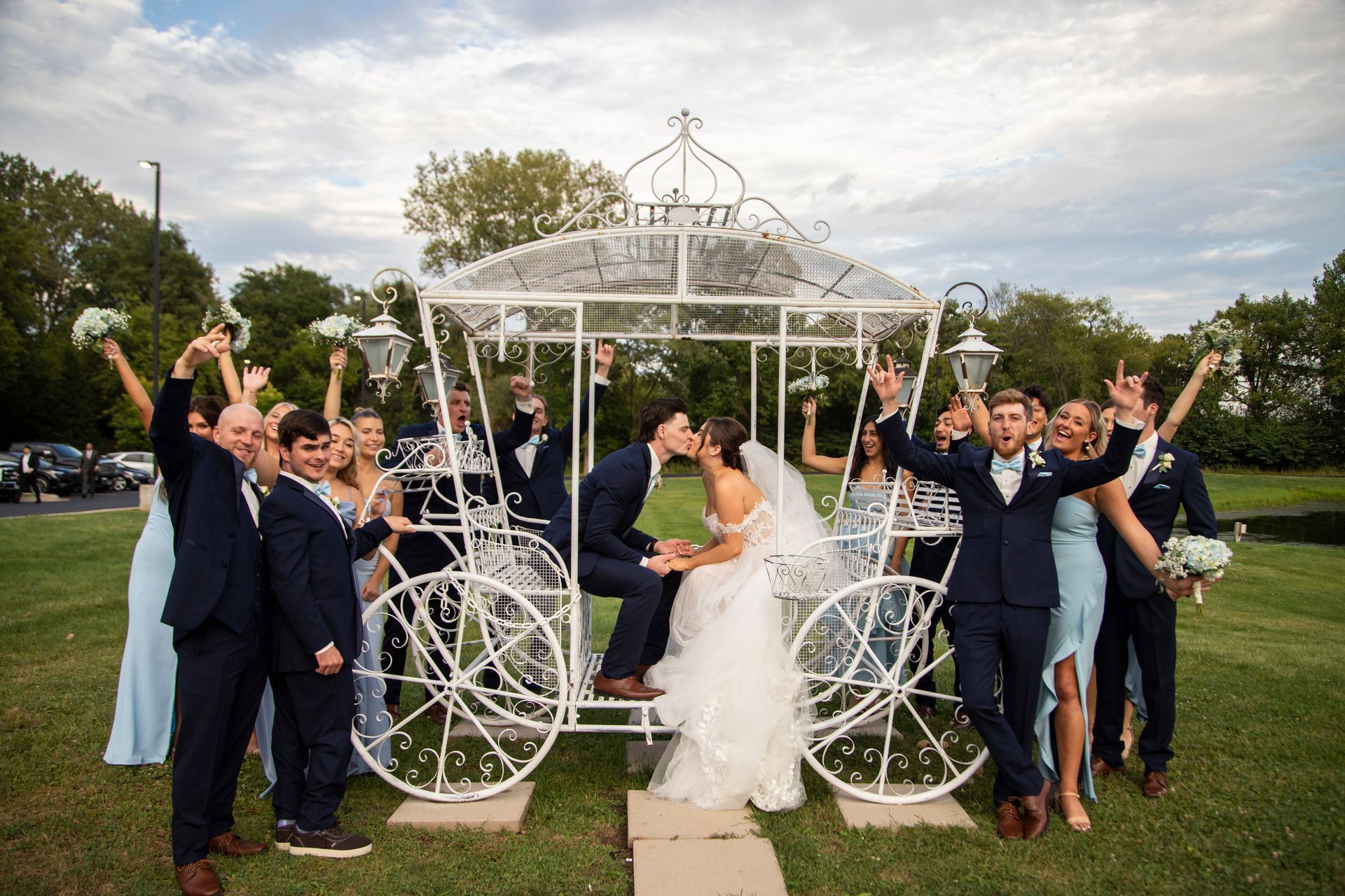 Wedding party celebrating, couple kissing in a white carriage. Others cheering, raising arms on grassy lawn.