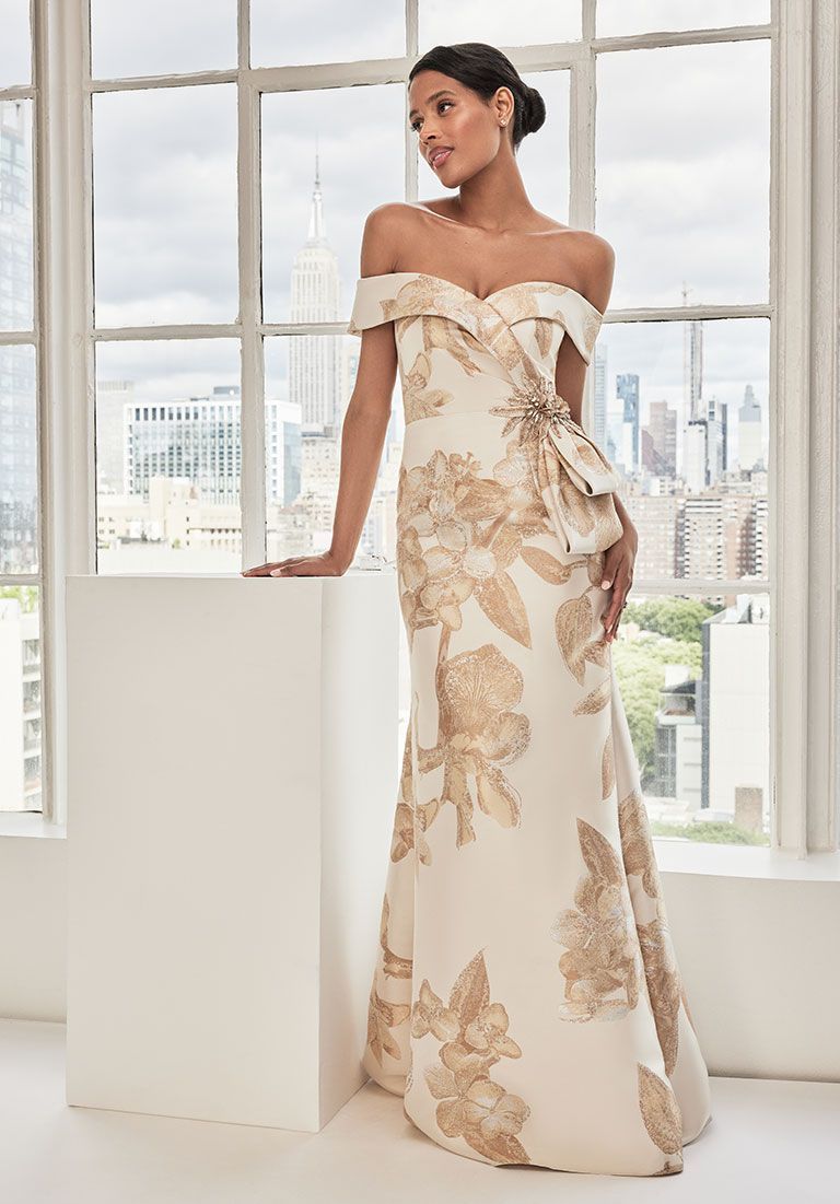 Woman wearing an off-the-shoulder, cream and gold floral-patterned gown, standing near a window overlooking a cityscape.