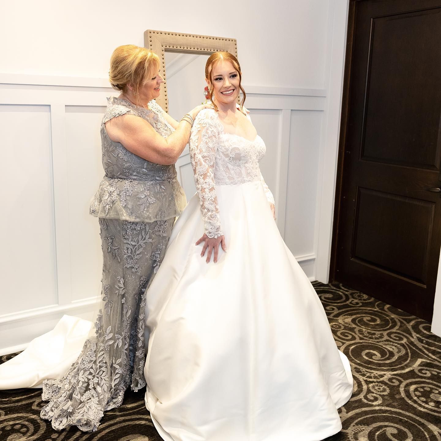 Woman in wedding dress, being helped by another woman in a silver gown, indoors near a mirror and door.