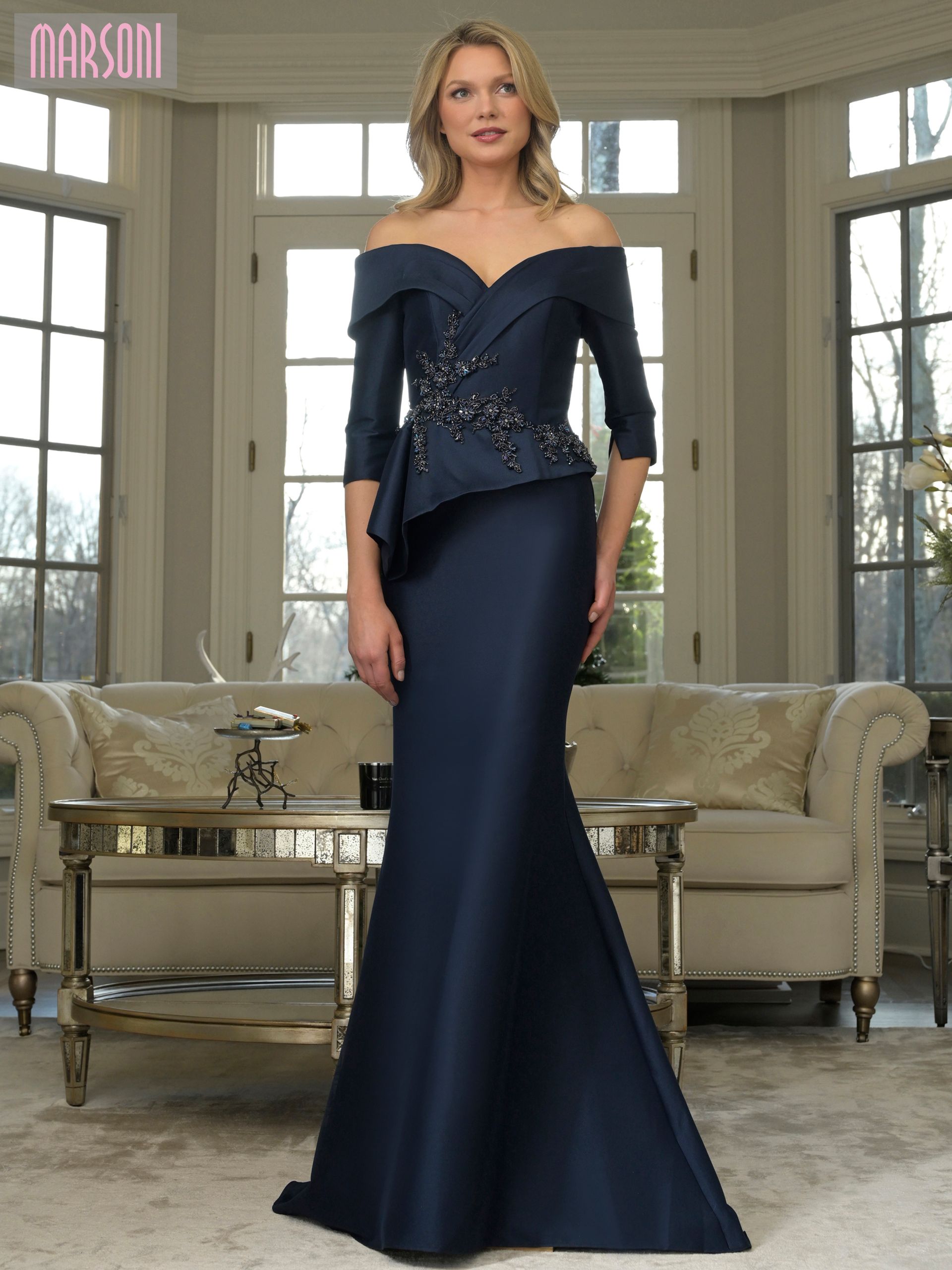 Woman in a navy off-the-shoulder formal gown, standing in a well-lit living room.