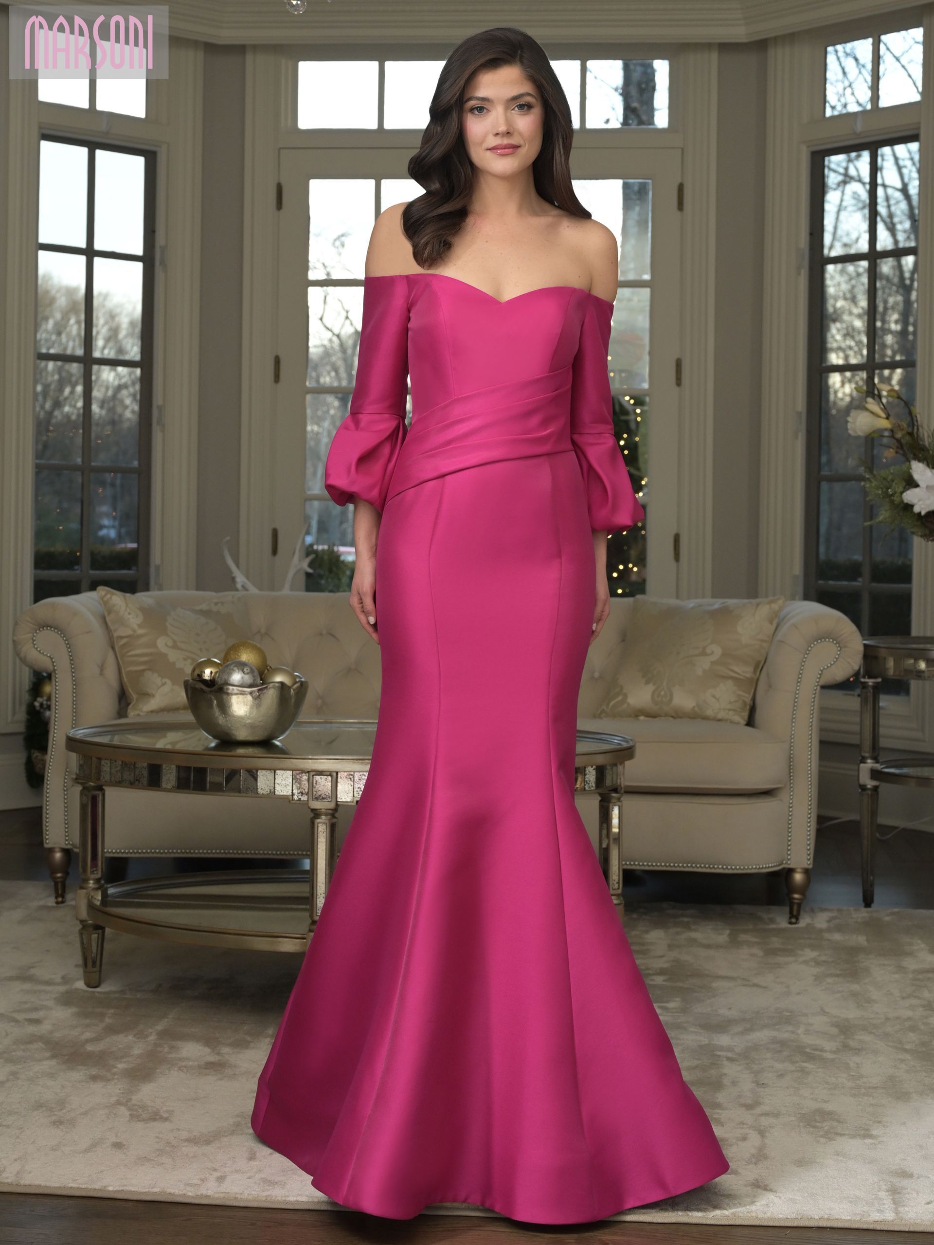 Woman in a magenta off-the-shoulder gown stands indoors. Dress has long sleeves and a mermaid skirt.