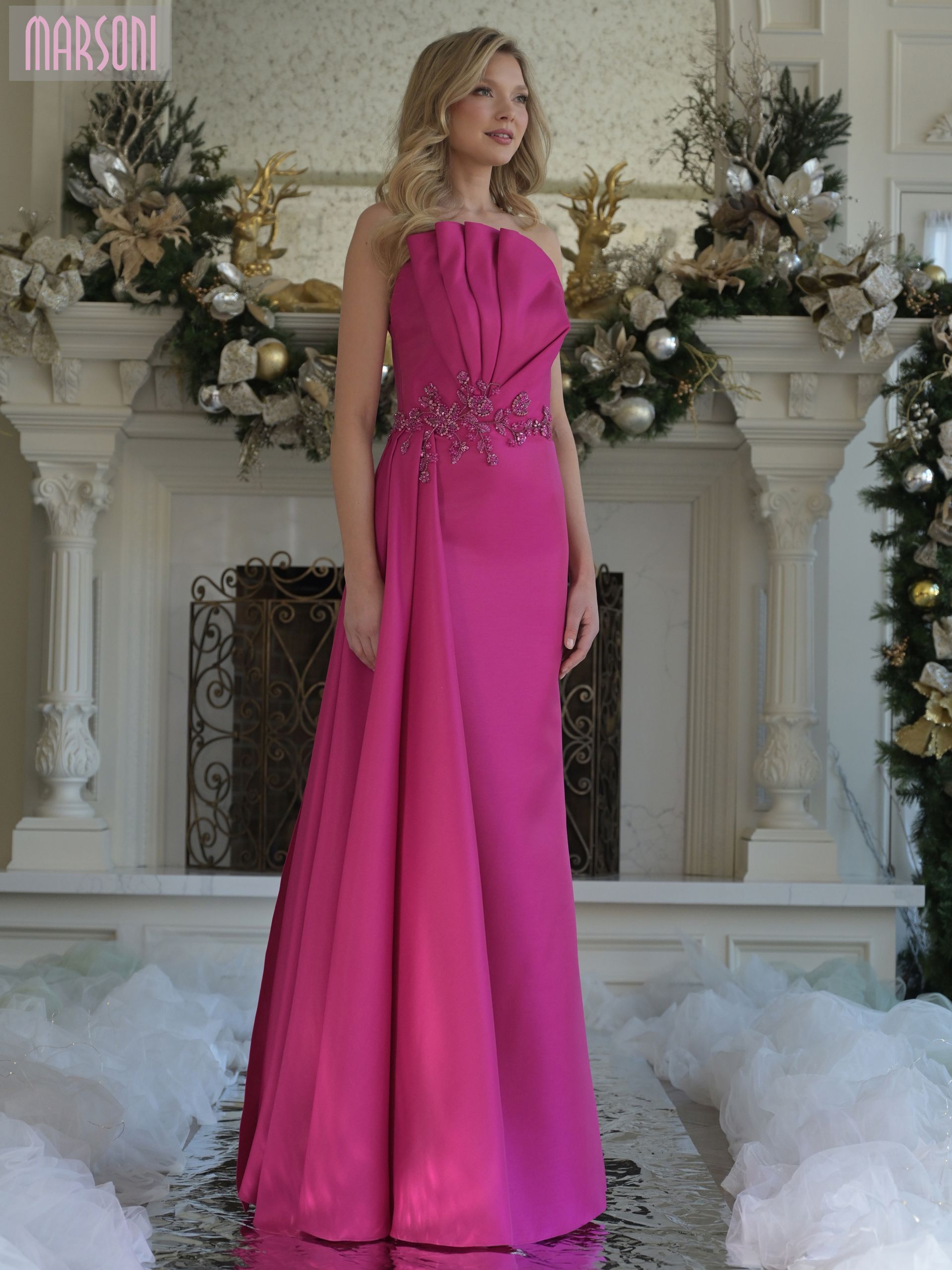 Woman in a magenta off-the-shoulder gown stands indoors. Dress has long sleeves and a mermaid skirt.