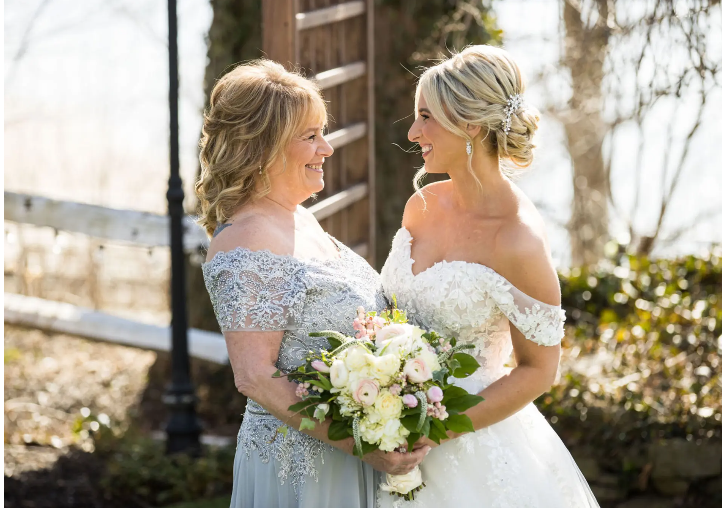 Bride and woman in lace embrace, smiling; holding flowers. Outdoor setting, sunny.