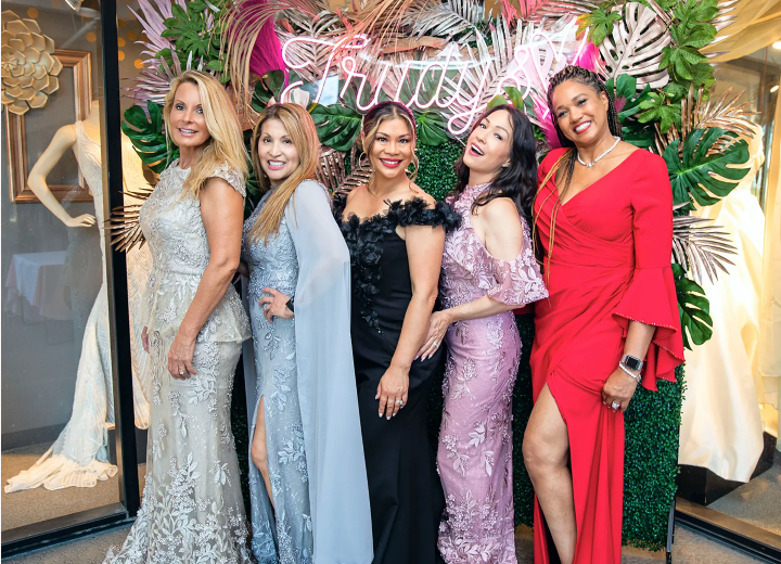 Five women in elegant gowns pose in front of a window display, smiling.