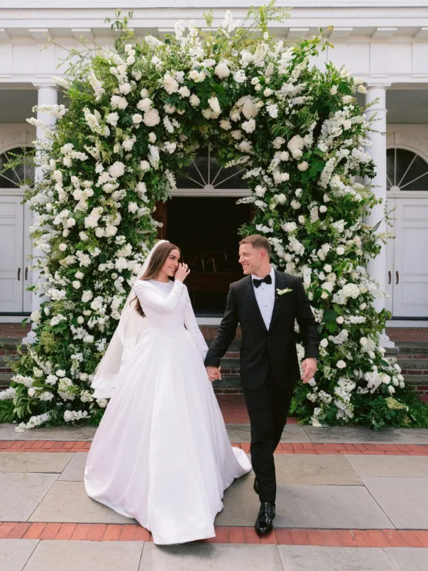 Newlyweds holding hands under a floral archway. Bride in white gown, groom in black tuxedo, walking out of a building.