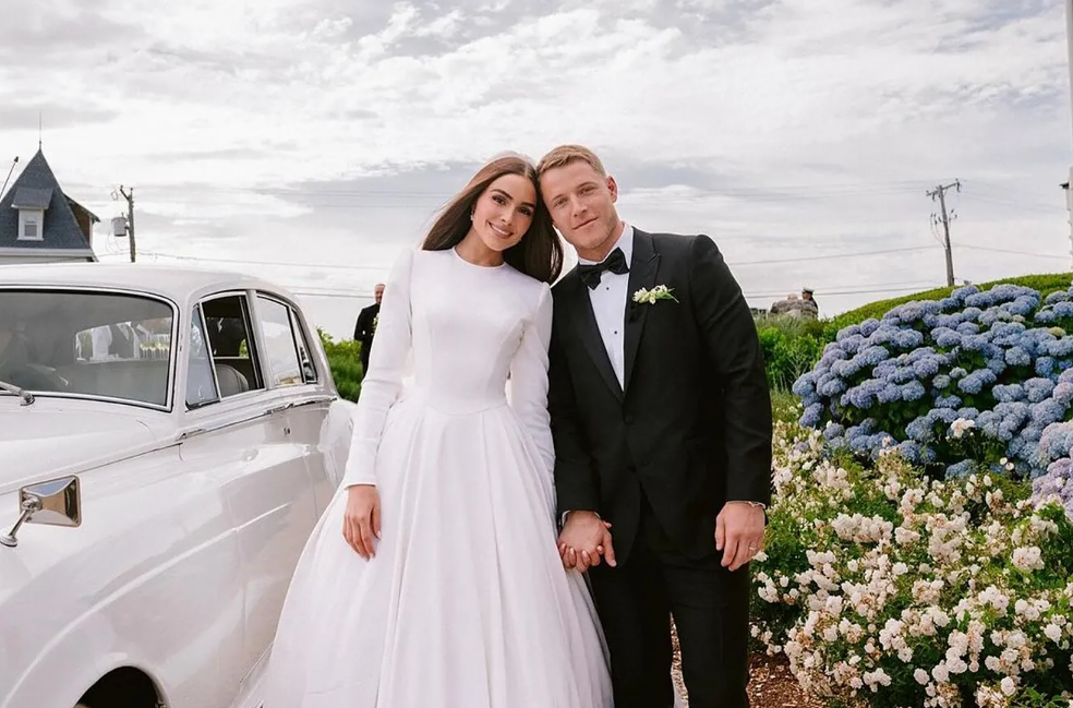 Newlyweds holding hands beside a white vintage car. The bride wears a white dress, the groom a black tuxedo.