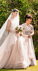 Bride in white dress and veil, holding bouquet, walks with woman in formal gown outdoors.
