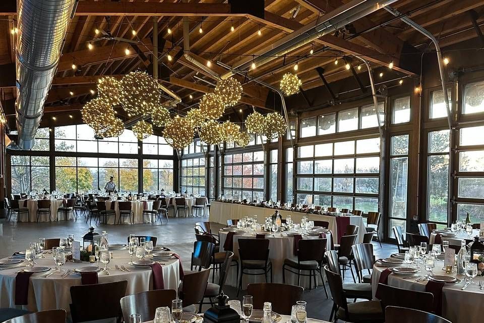 Inside a venue: tables set for a celebration, with round tables, dark chairs, and large windows looking out at fall foliage.