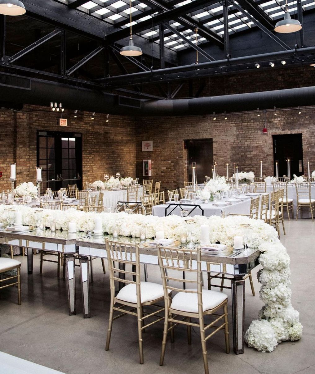 Wedding reception hall with long tables, white flowers, gold chairs, exposed brick walls, and skylights.