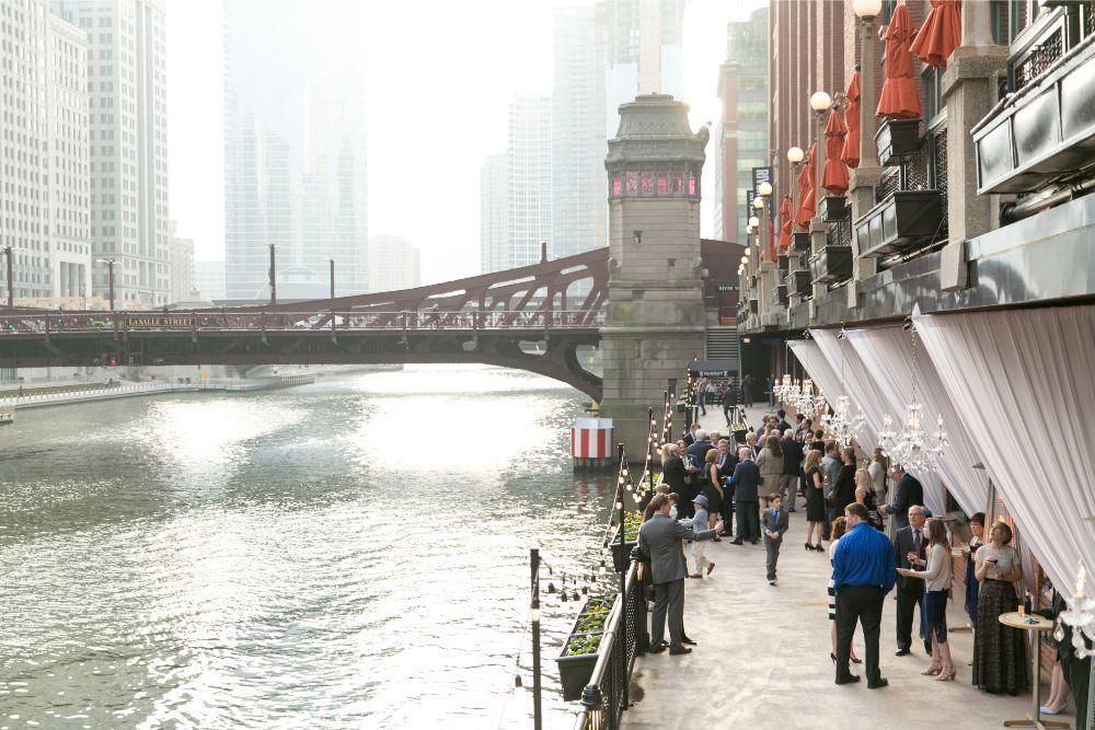 Chicago Riverwalk with crowd near buildings and bridge. Water, buildings, and overcast sky.