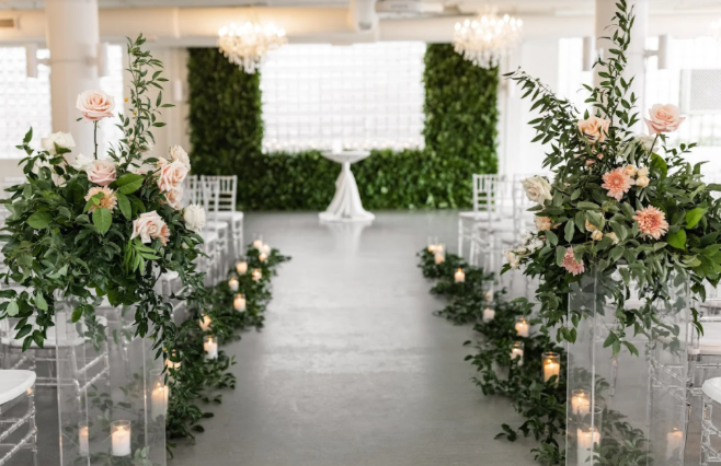 Wedding aisle with floral arrangements and candles. Greenery backdrop, white chairs.