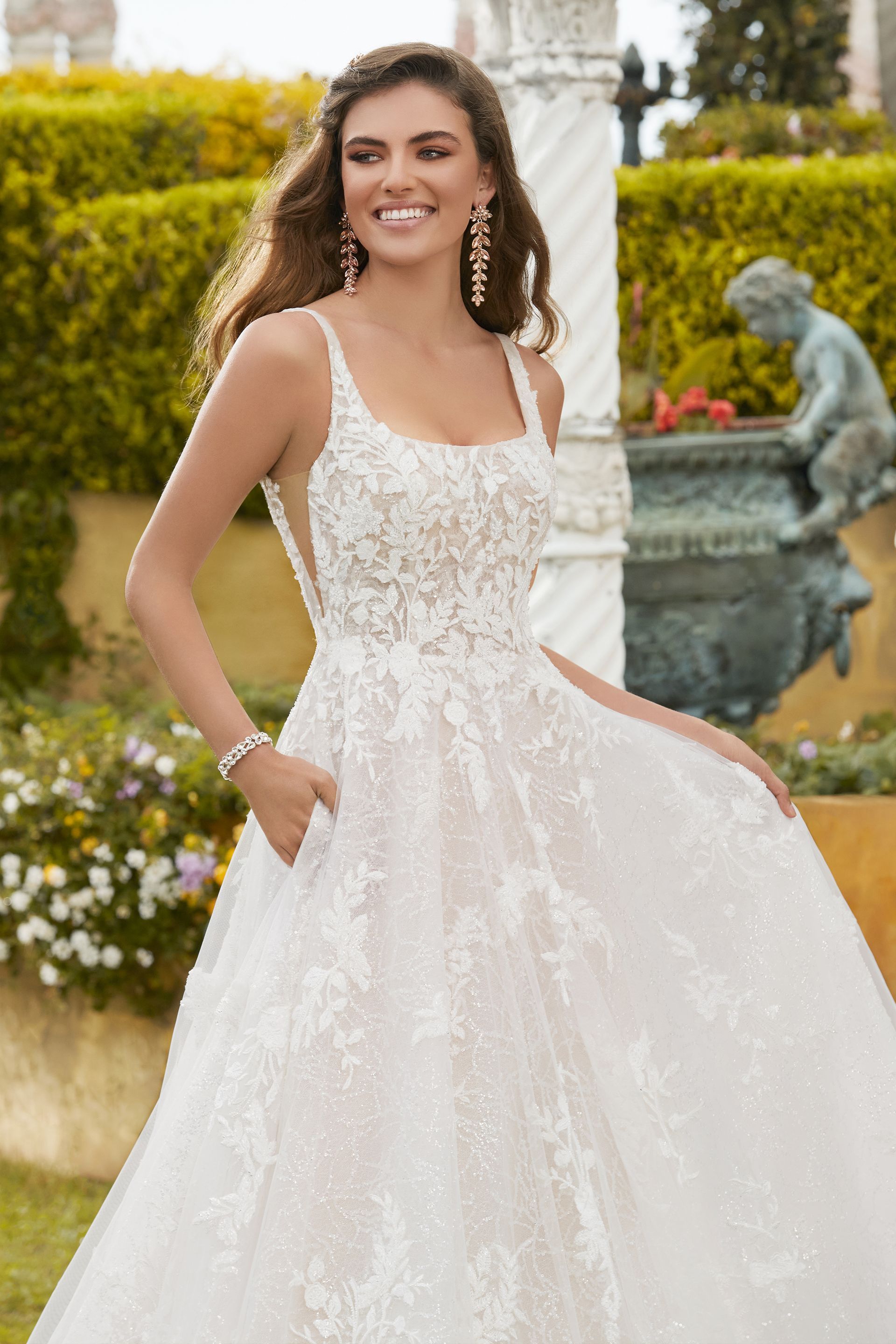 Woman in a white wedding gown smiles outdoors. She stands near a garden with a fountain.
