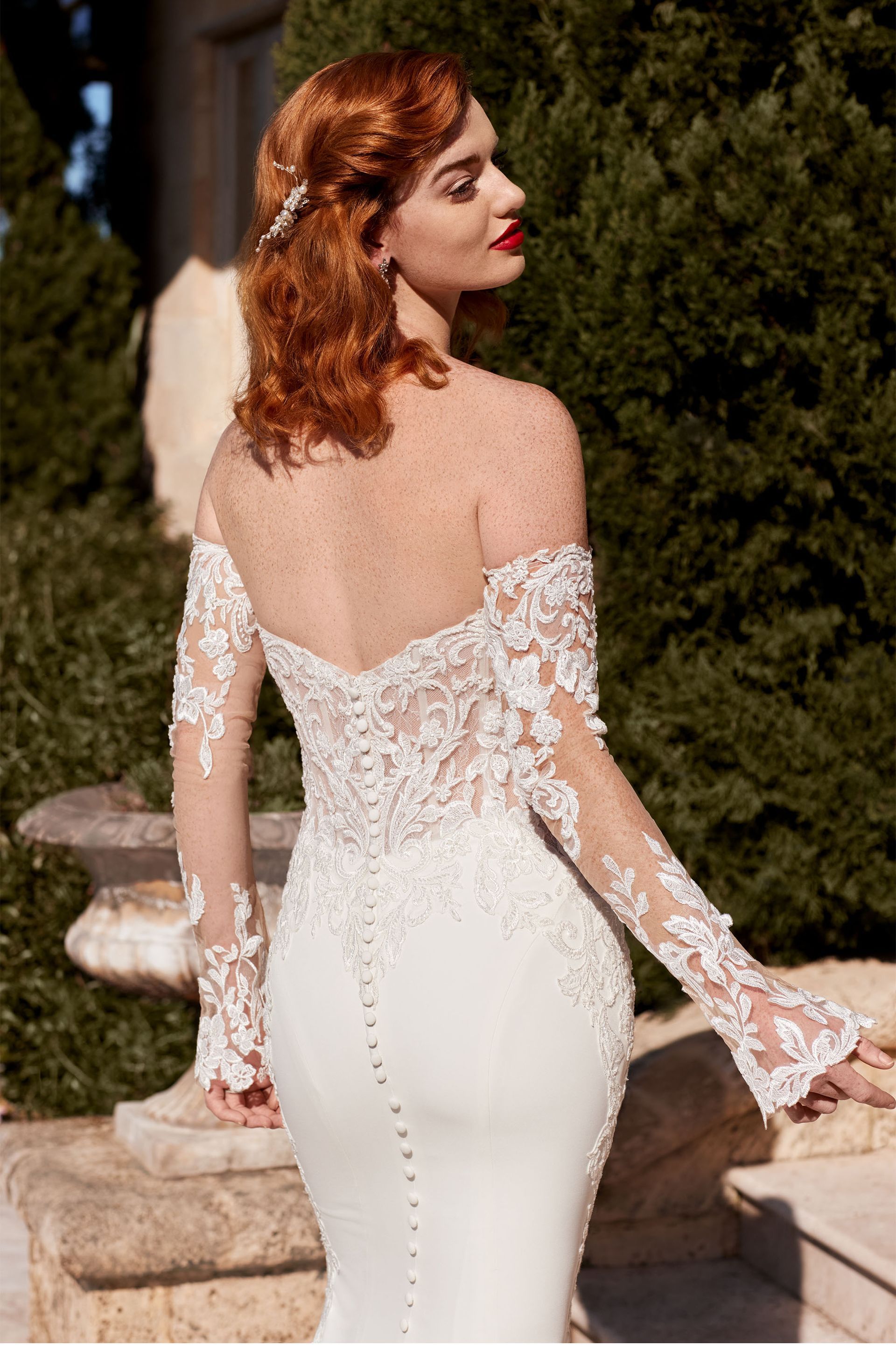 Woman in white, off-the-shoulder wedding gown with lace sleeves and a flowing train, standing outdoors.