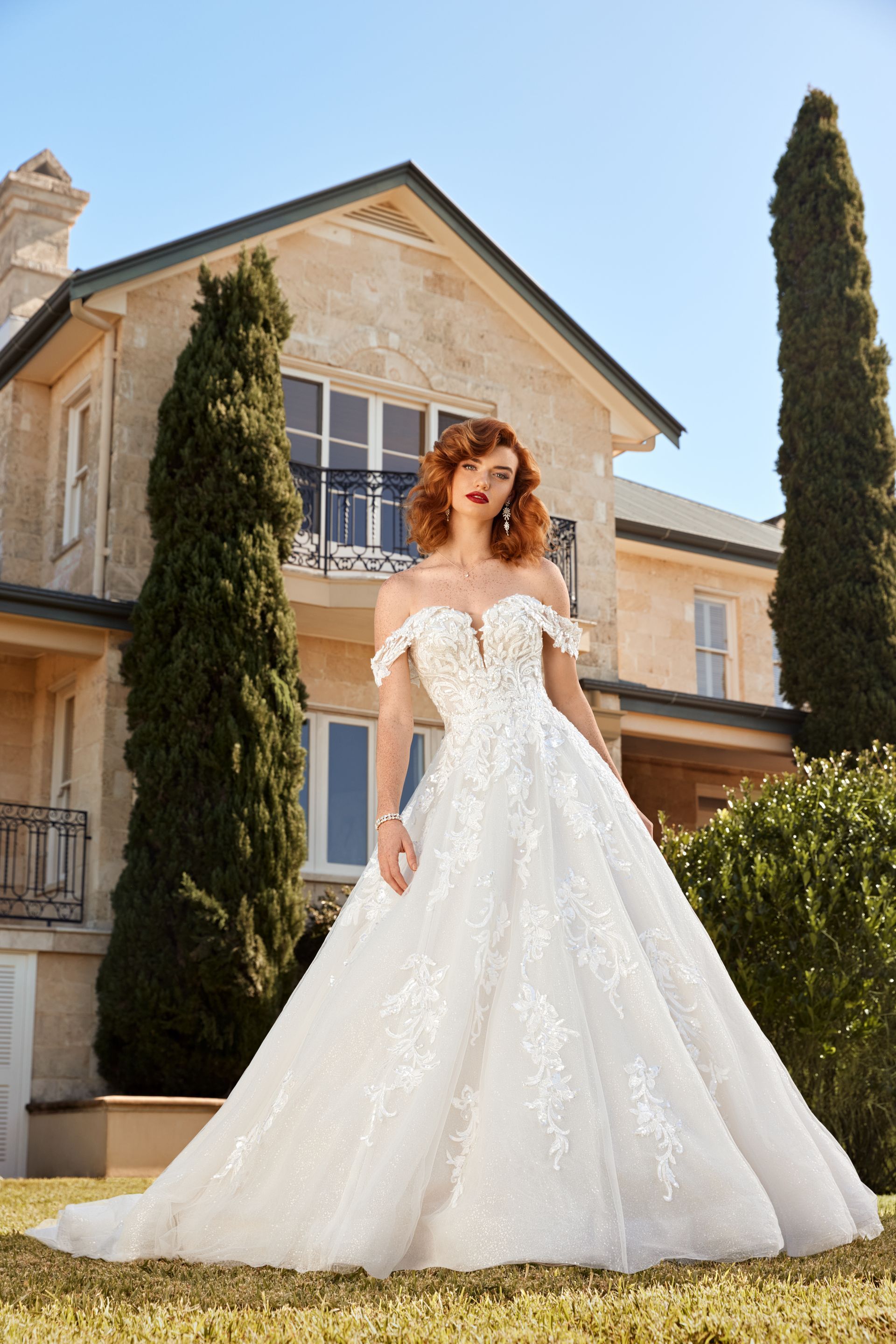 Woman in off-the-shoulder wedding gown stands on lawn in front of a house.