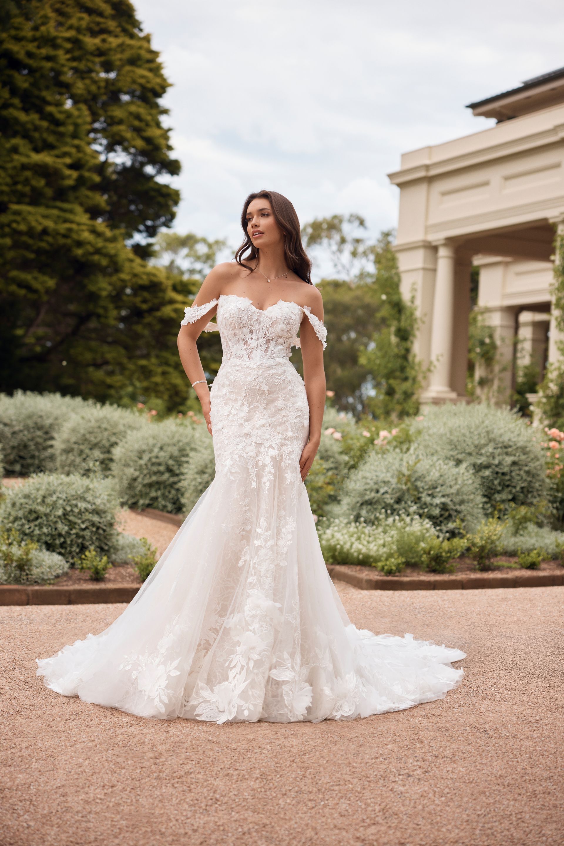 Woman in white, off-shoulder wedding dress posing outdoors in front of a garden and building.