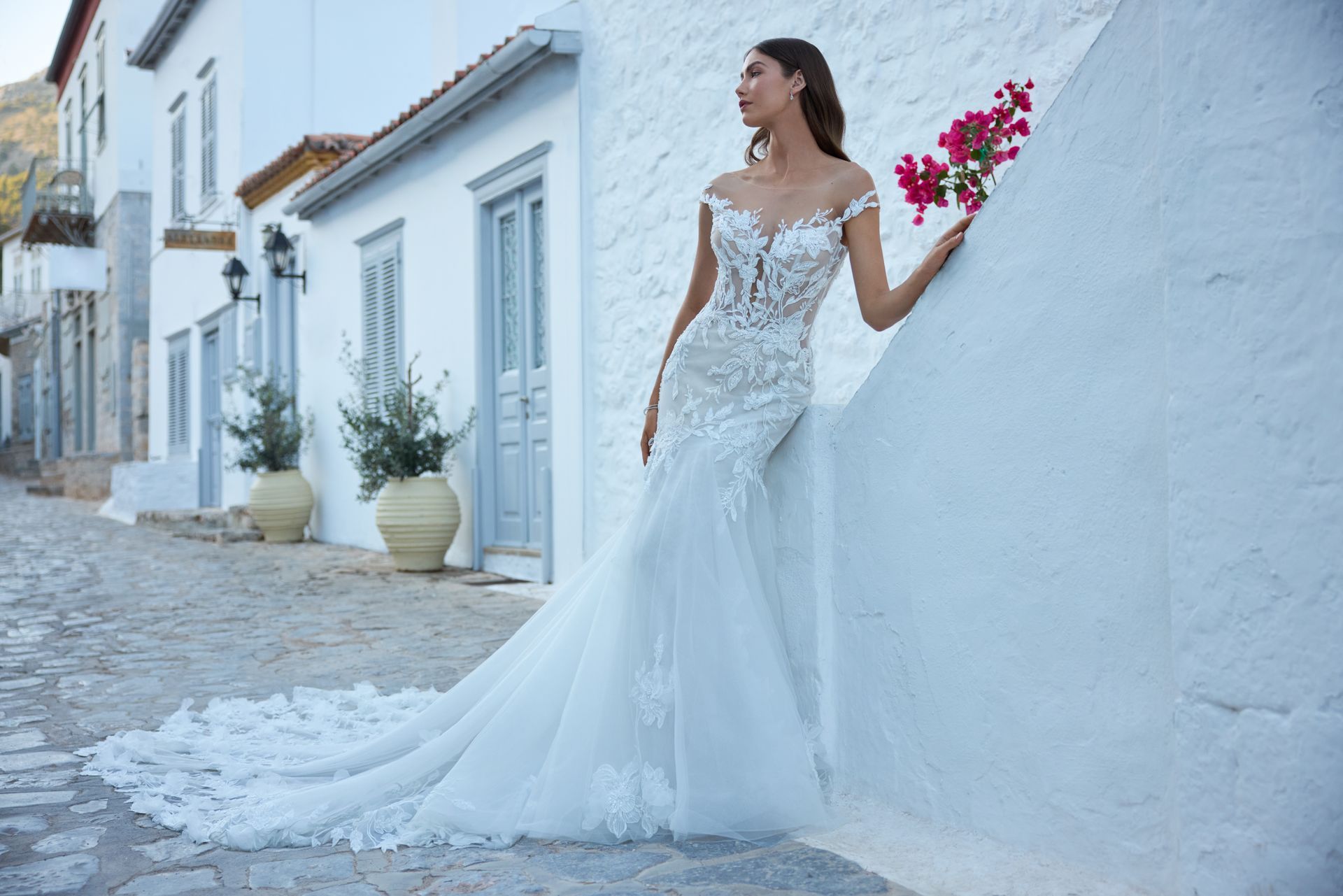Woman in a white wedding dress with a long train, holding flowers, leaning against a white wall.
